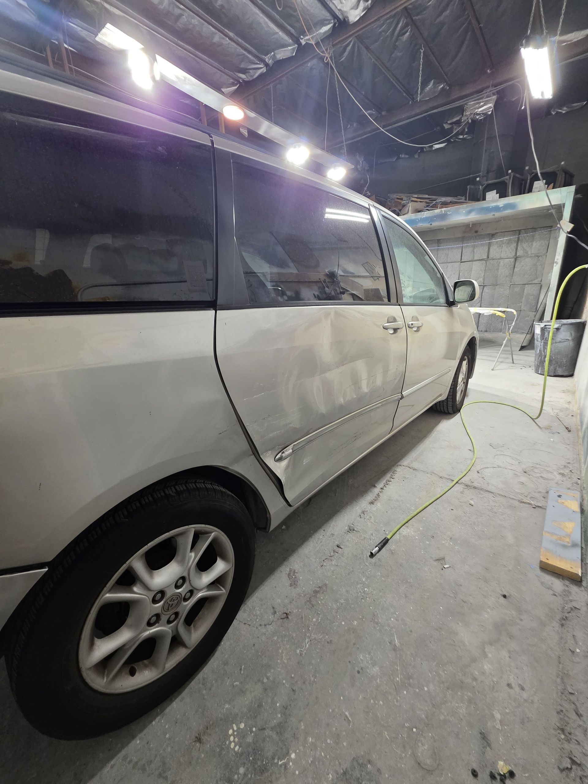 Silver minivan in a dim auto body shop with a dented side panel and wheel visible