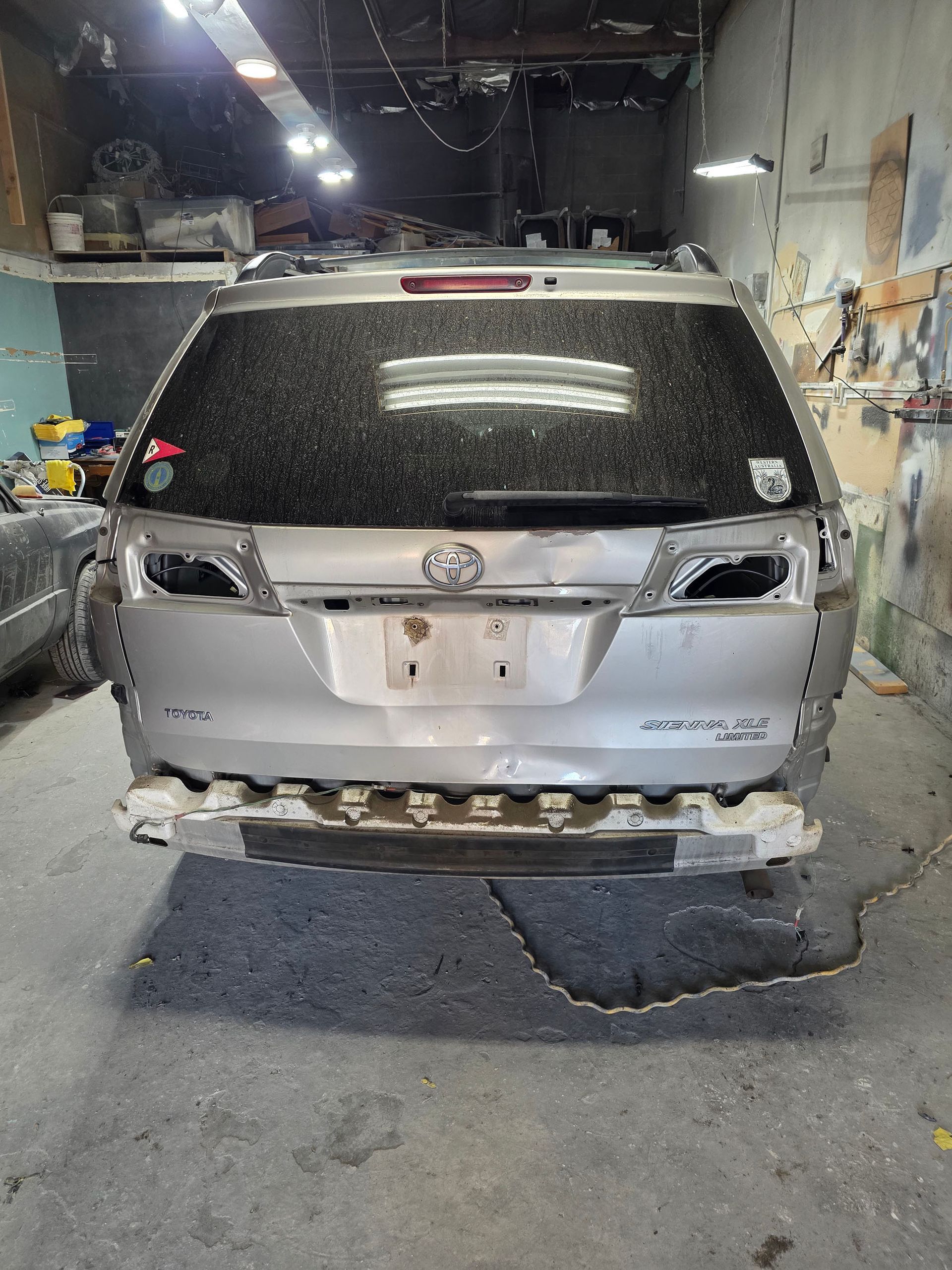 Damaged silver SUV in a garage, rear bumper removed and rear window shattered.