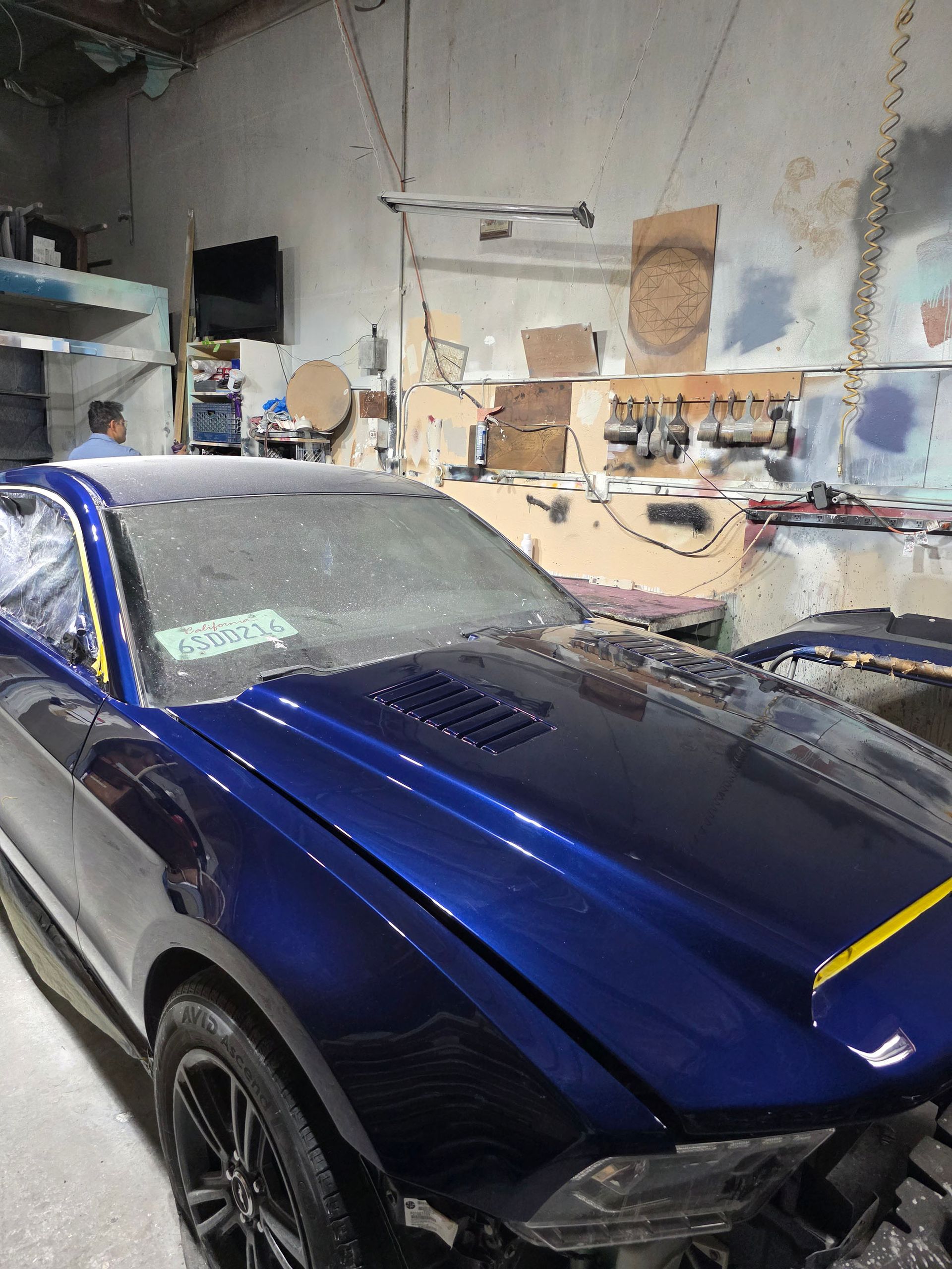 Blue sports car in a workshop, with tools and supplies on shelves in the background.