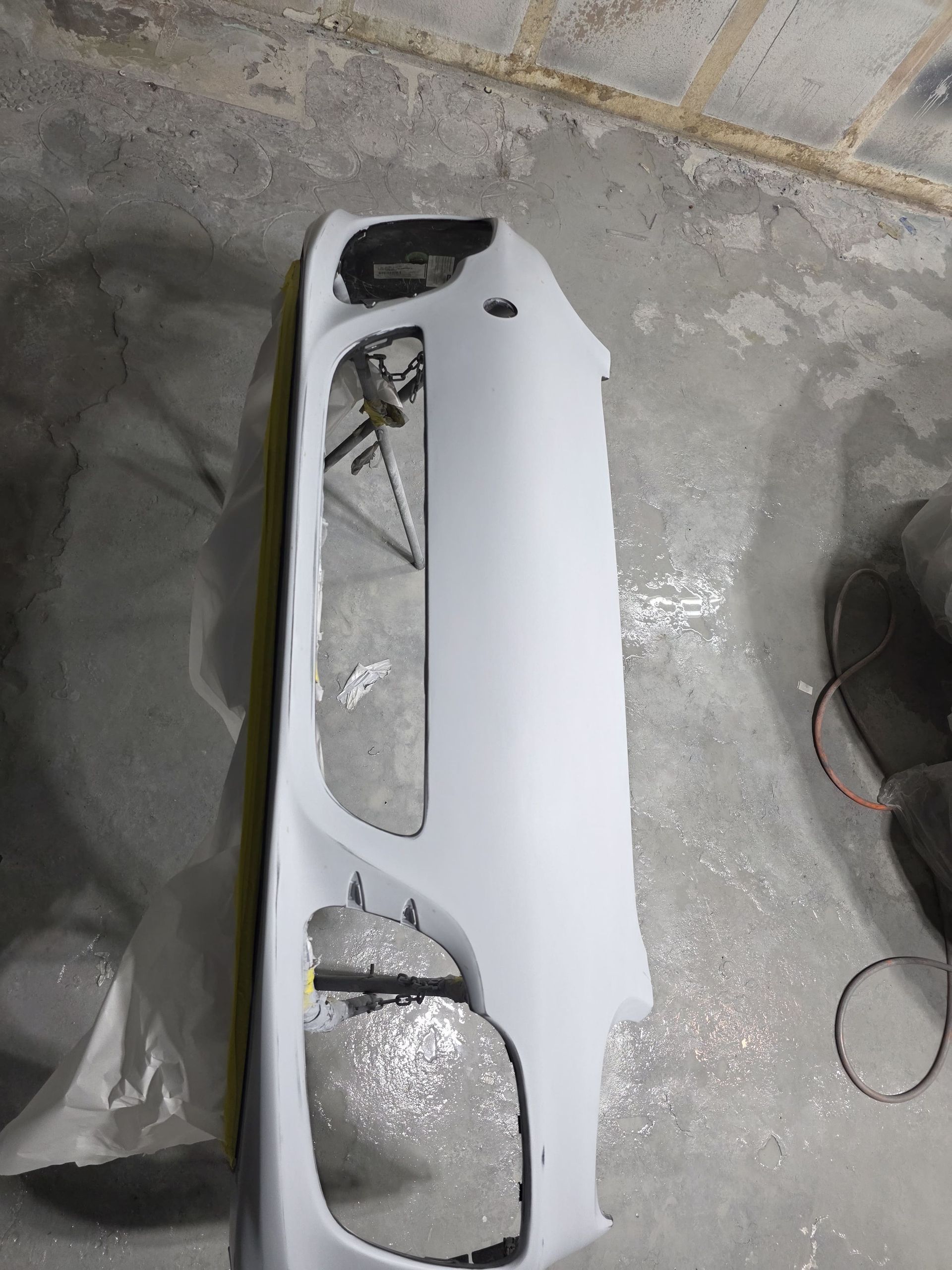 White car bumper cover lying on a concrete floor, with cutout openings and visible damage.
