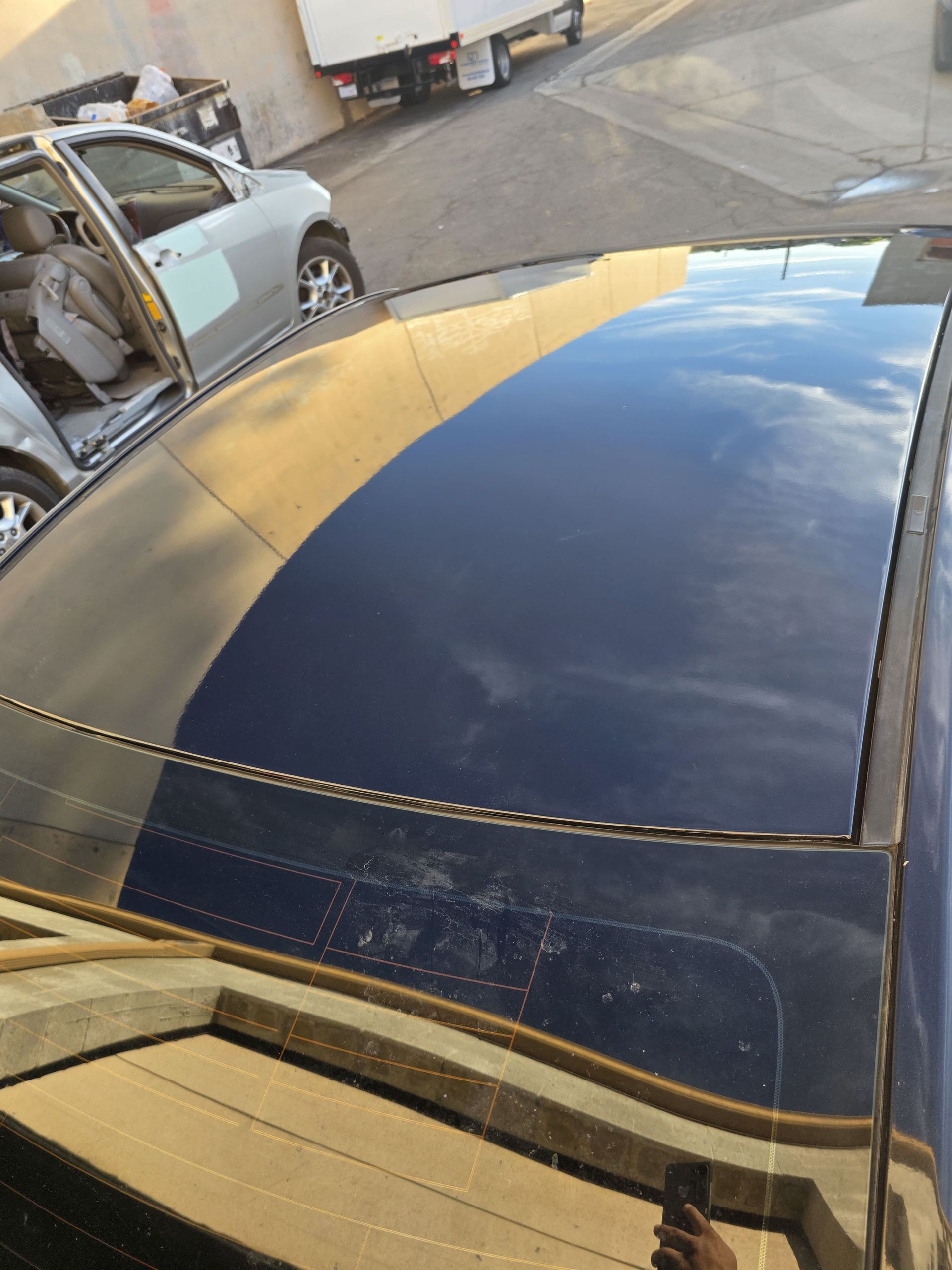 Black car roof with a large glossy panel reflecting the blue sky in a parking lot.