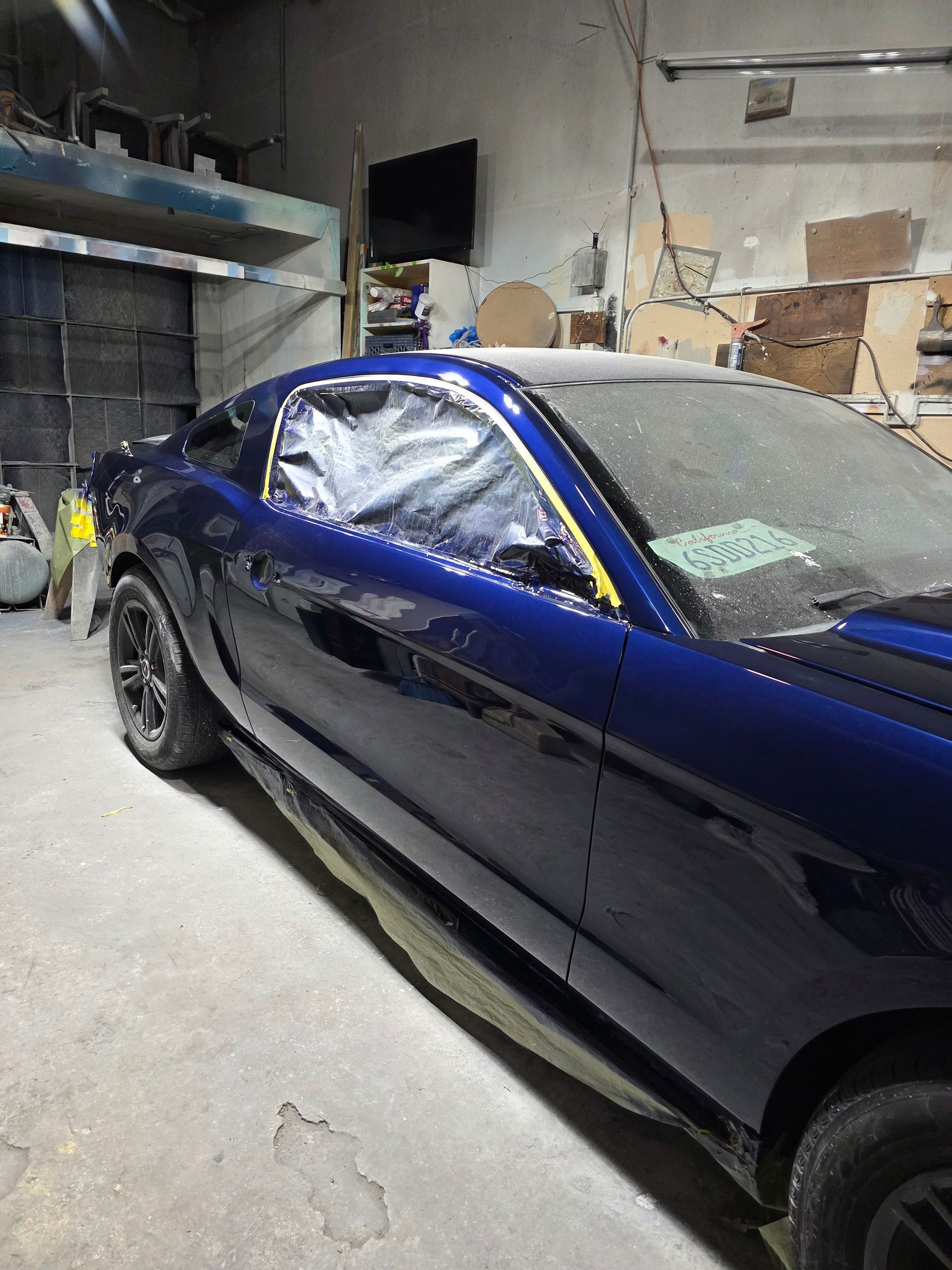Blue sports car in a workshop, partially wrapped in clear plastic with walls and tools in the background