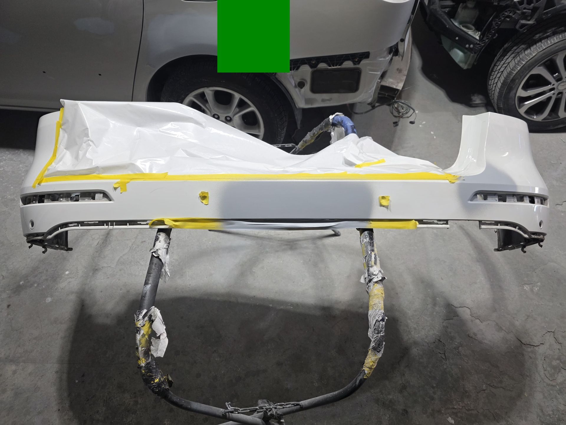 Damaged white car bumper on a stand in a garage, with yellow tape and exposed inner parts.