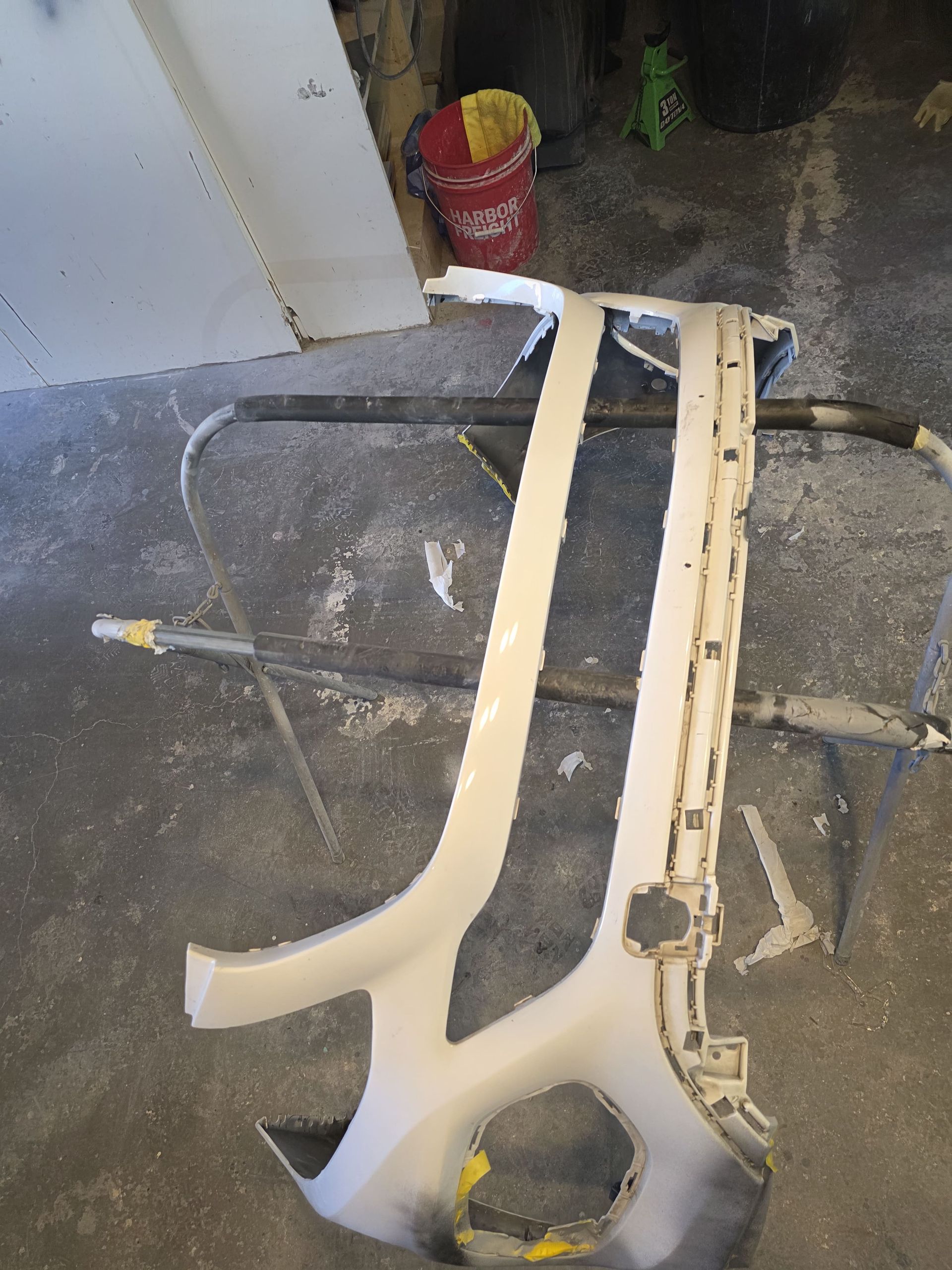 White car bumper parts leaning on a sawhorse in a workshop or garage, with tools and a bucket nearby