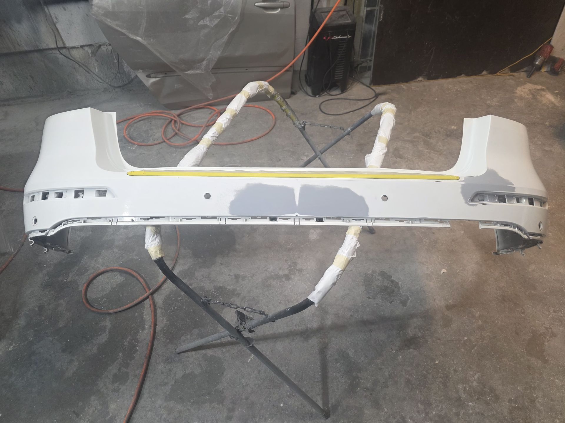 White car bumper being painted in a workshop, mounted on a stand with taped edges and gray primer patches.