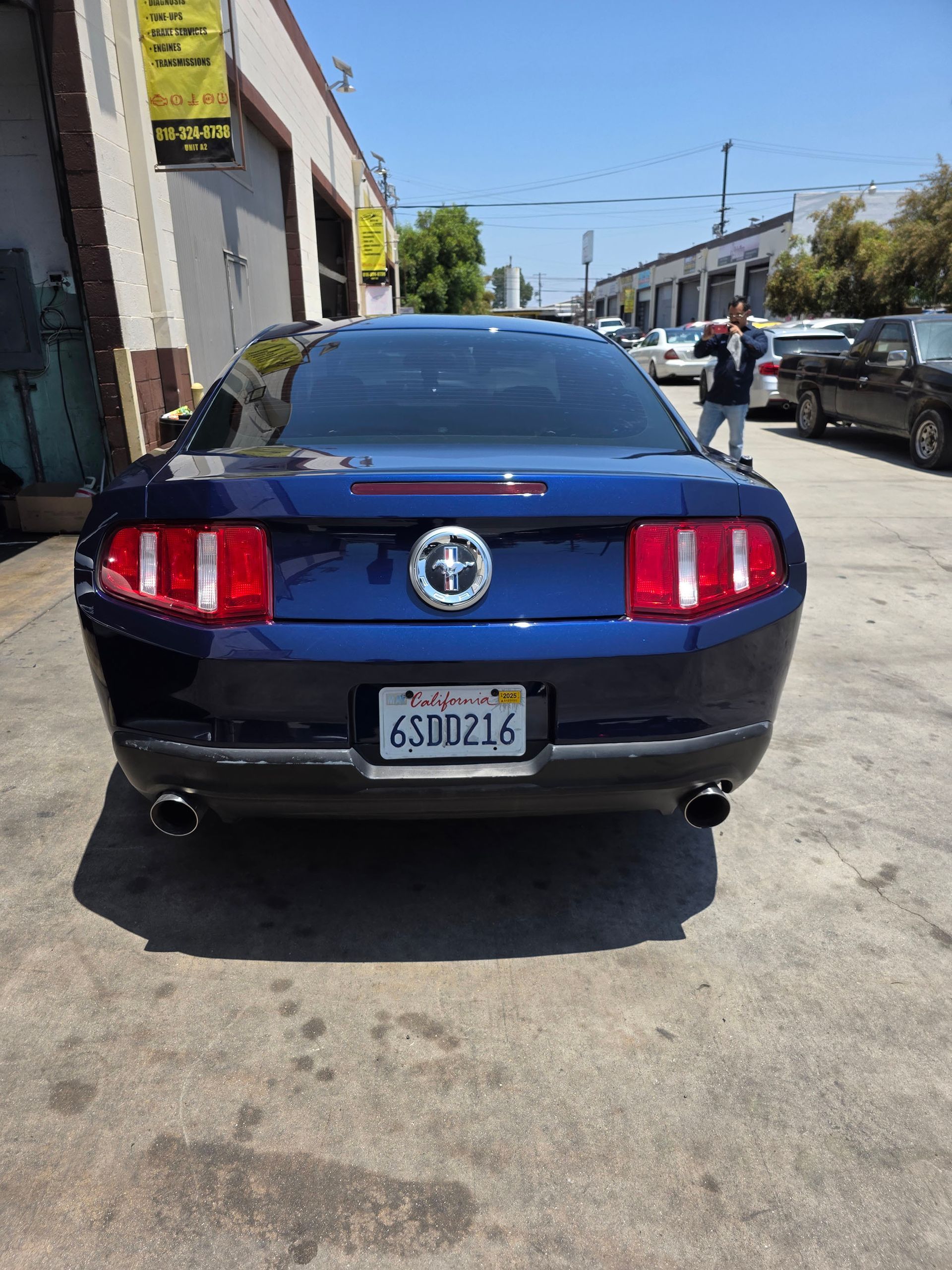 Rear view of a dark blue Ford Mustang parked on a sunny street beside a building.