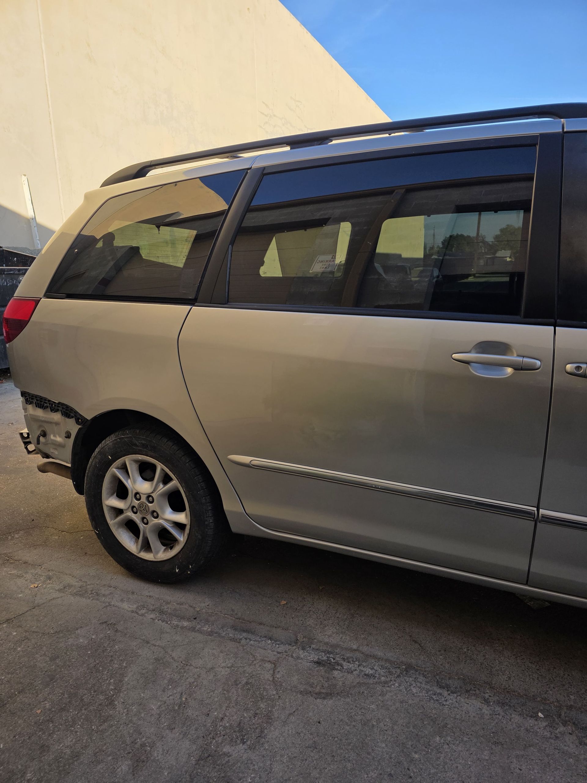 Silver minivan parked beside a beige wall on a paved street, viewed from the side.