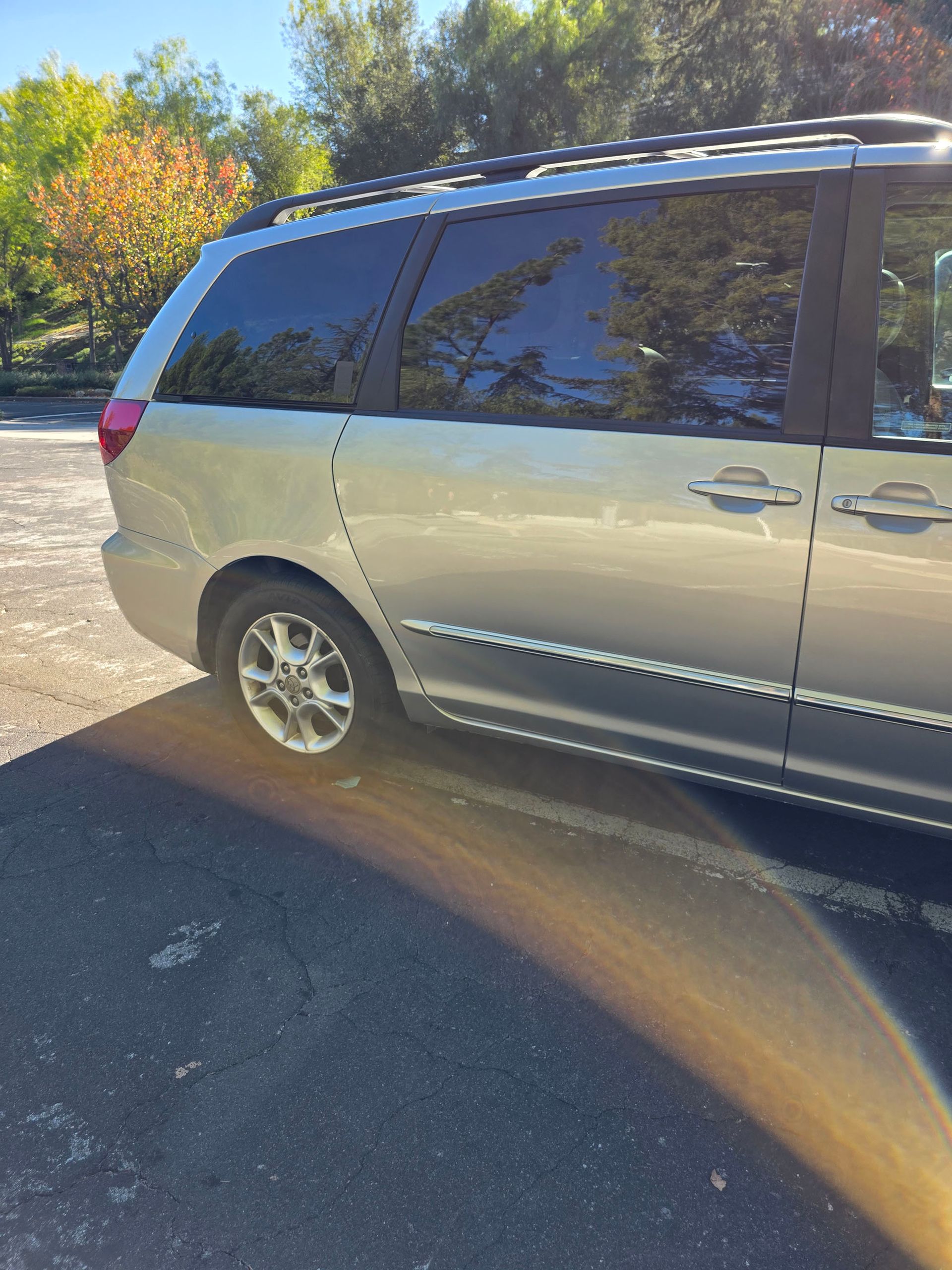 Silver minivan parked on a sunlit driveway beside autumn trees, side view.