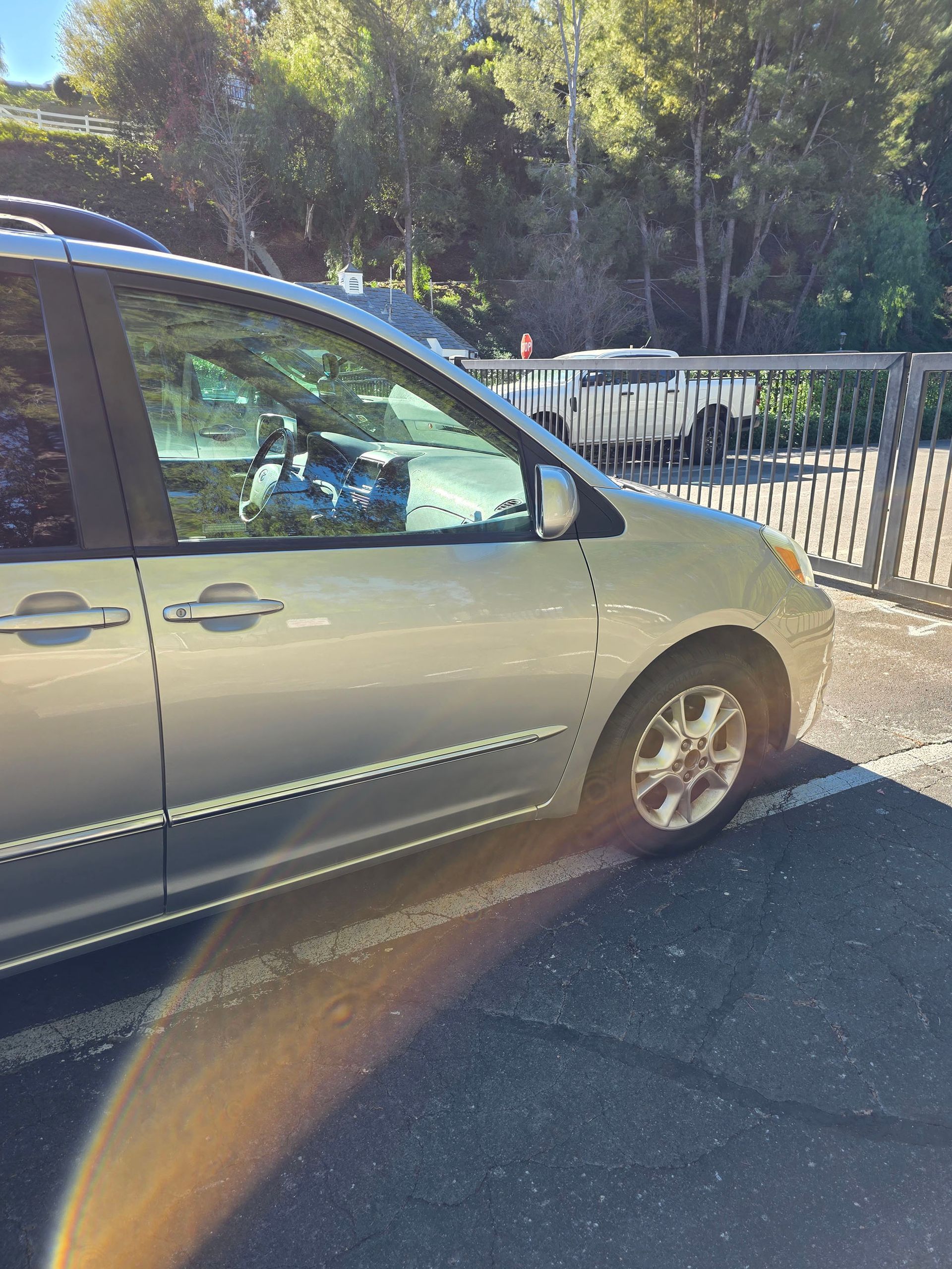 Silver minivan parked beside a metal fence on a sunny road, with trees in the background