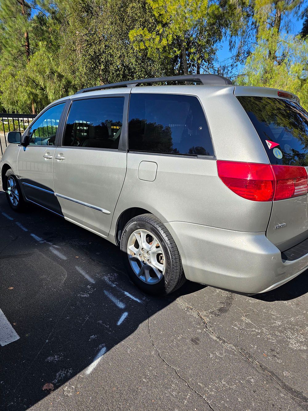Silver minivan parked on a sunny road beside trees, rear side view