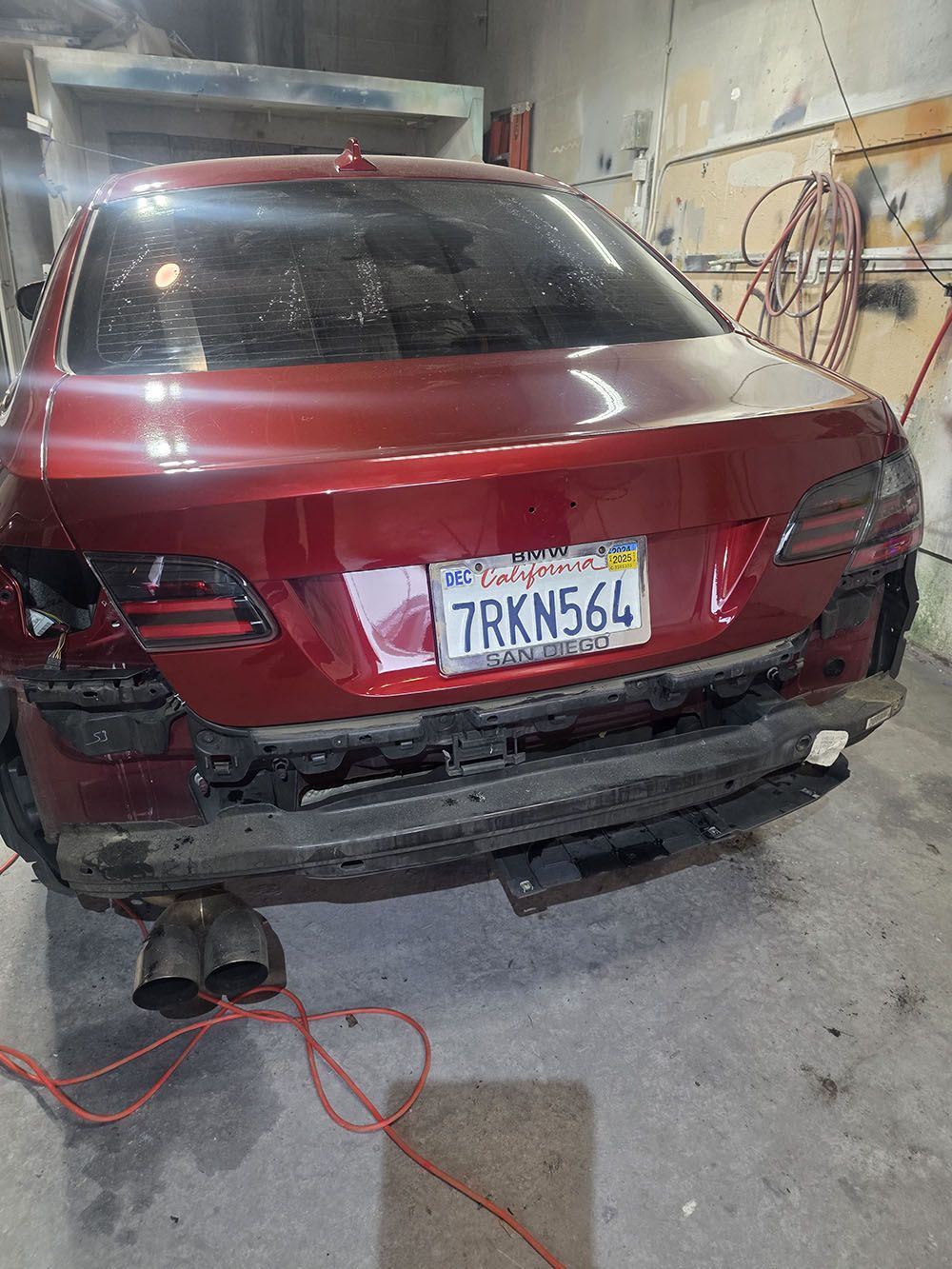 Red sedan in a repair shop, rear bumper removed and license plate visible.