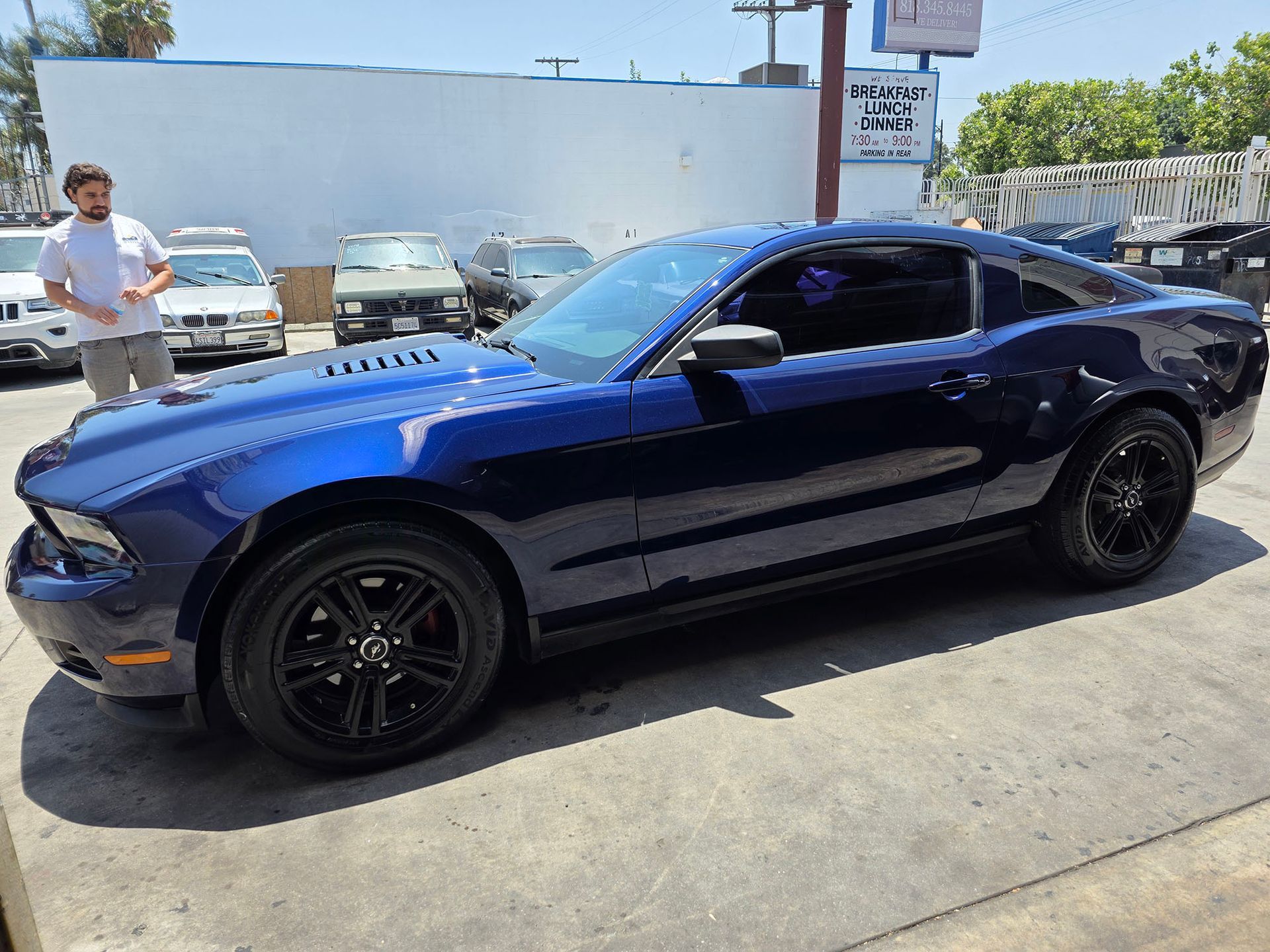 Blue sports car parked outdoors in a sunny lot, with people and other cars in the background