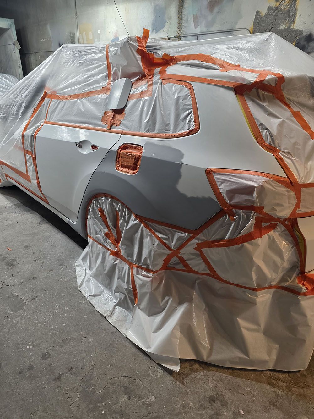 Car covered in white protective wrap and orange tape inside a garage or workshop