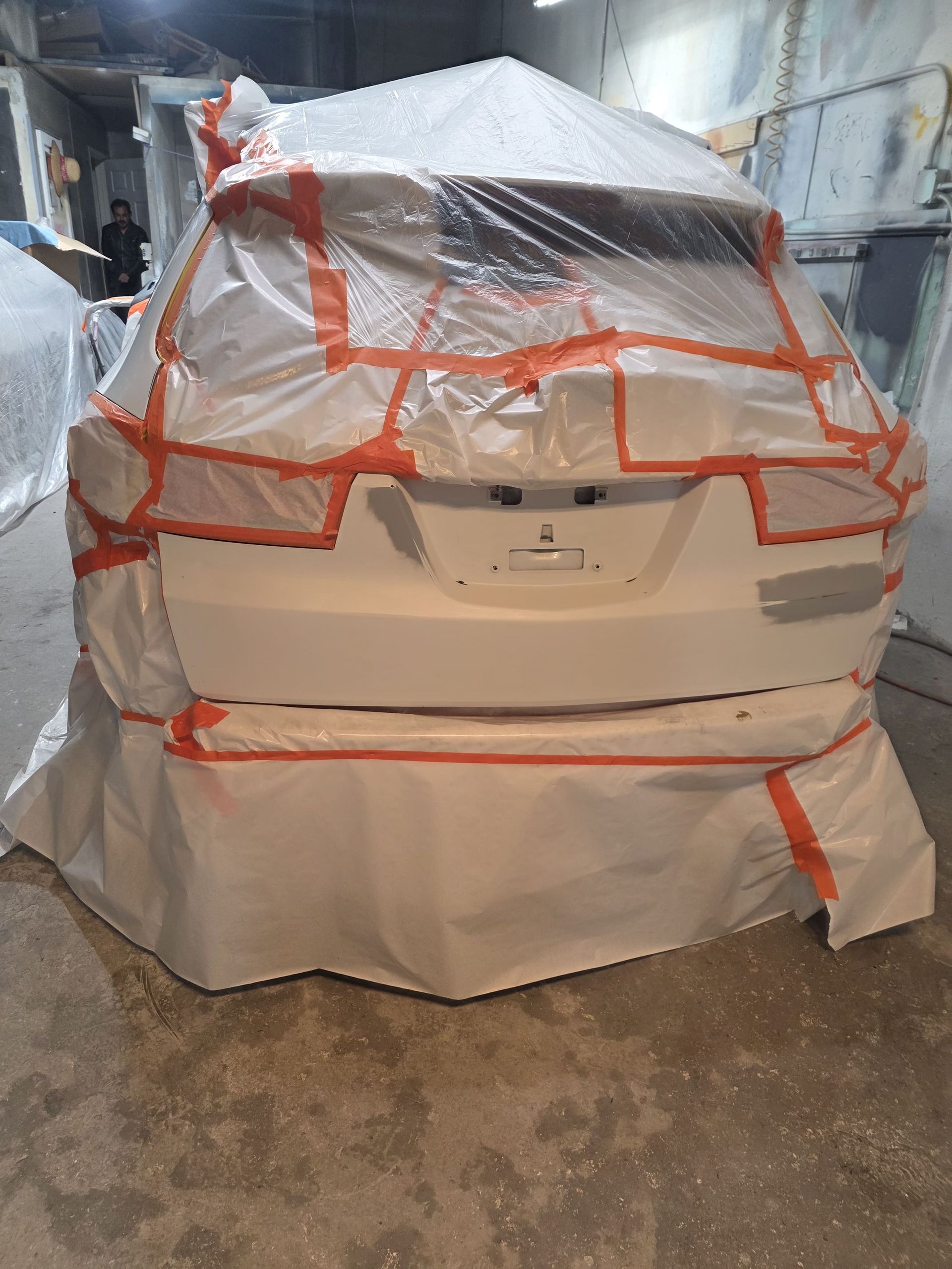 Car covered in white protective plastic with orange tape, parked indoors in a garage or storage area