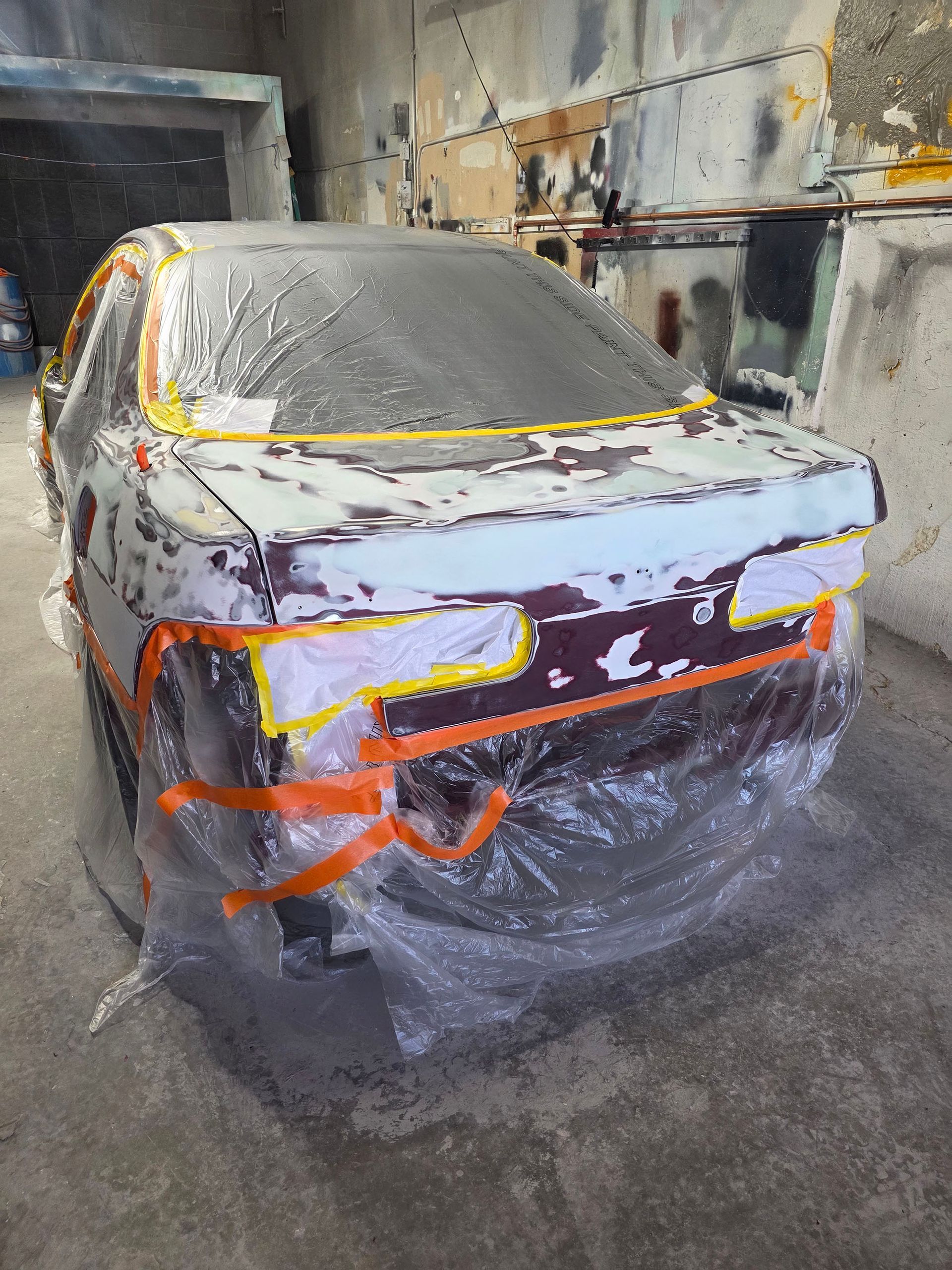 Sedan wrapped in plastic and masking tape in a paint booth, primed for bodywork and paint.