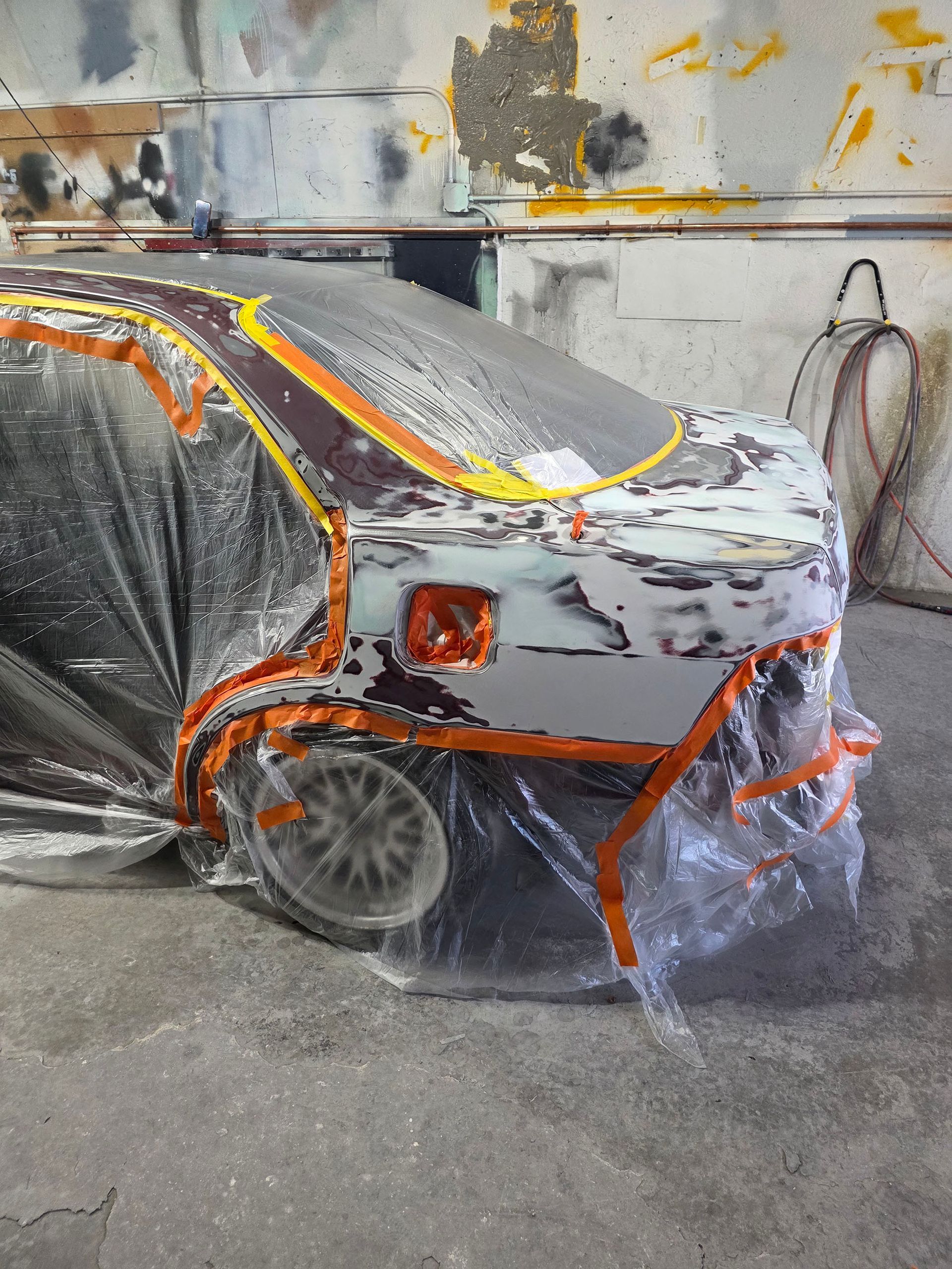 Car under repair, masked in plastic and tape, with gray primer and orange paint marks in a workshop.