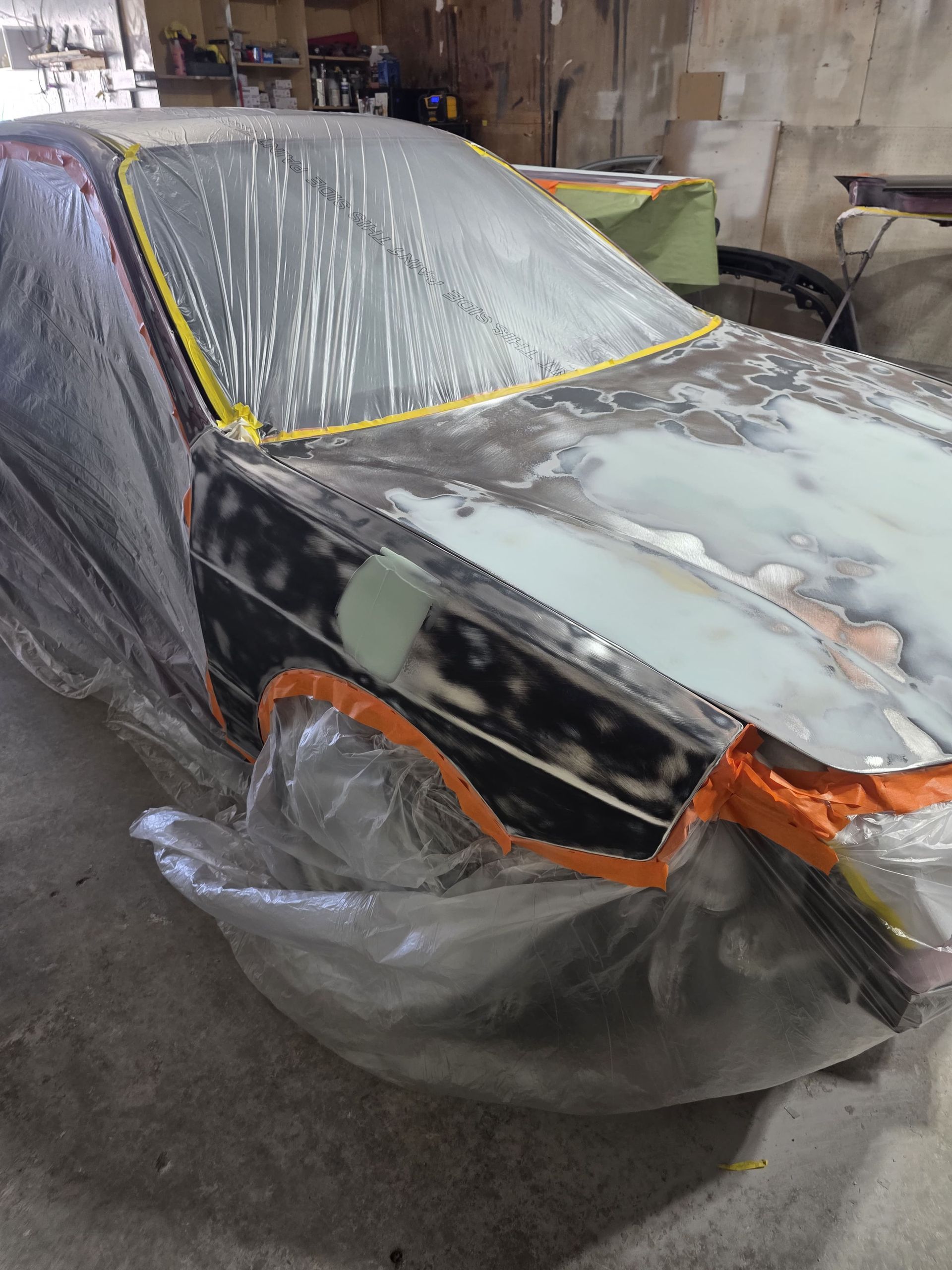 Car covered in plastic and masking tape in a workshop, with gray primer on the hood and body.