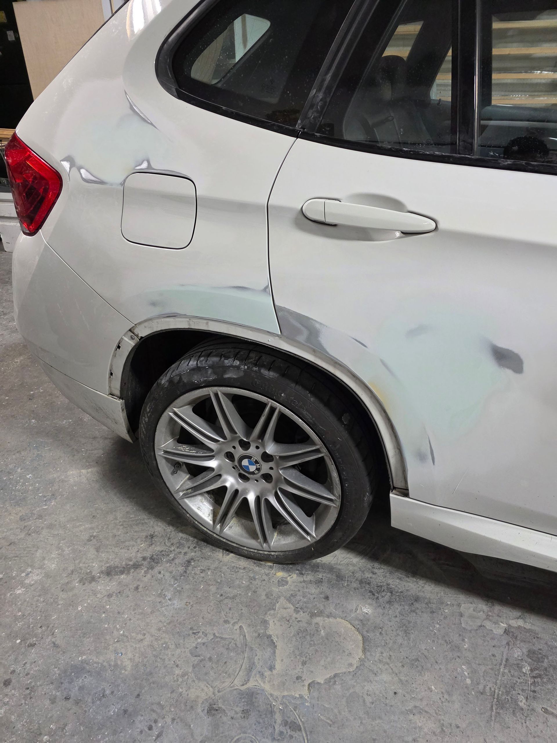 Rear side of a white SUV with a damaged rear wheel and scuffed body panels in a garage