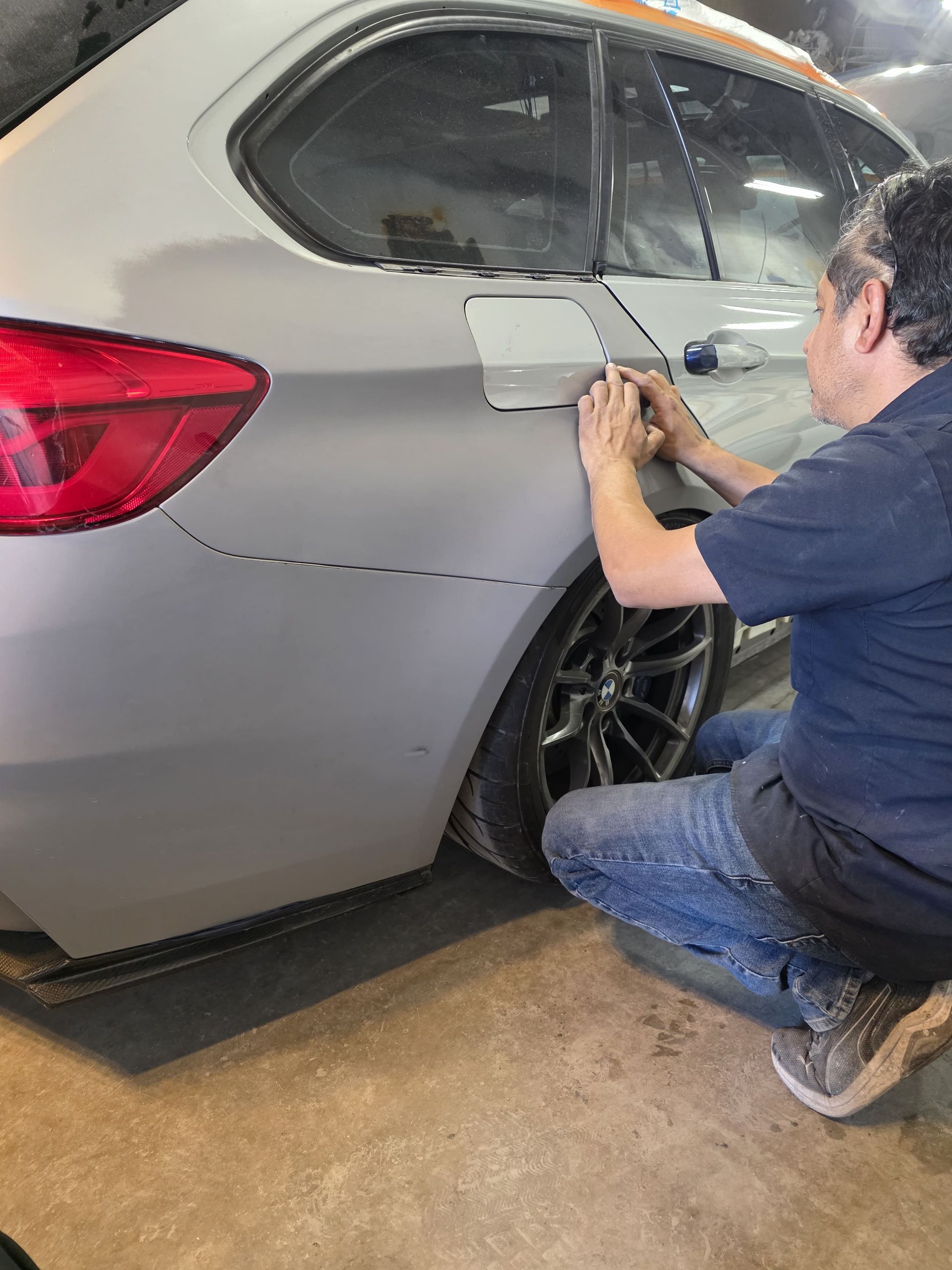 Person crouches beside a silver car, working on the rear door in a garage.