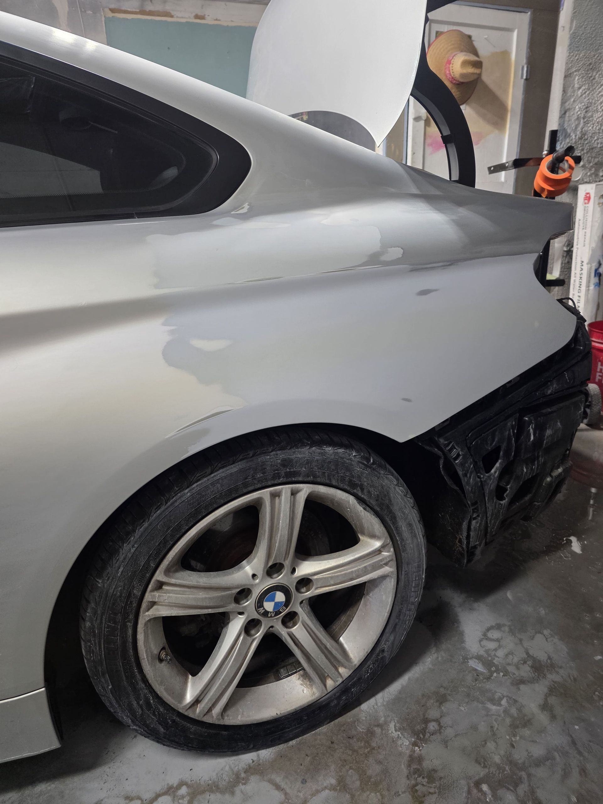 Silver BMW coupe in a workshop, rear wheel and bumper area showing damage or repair work.