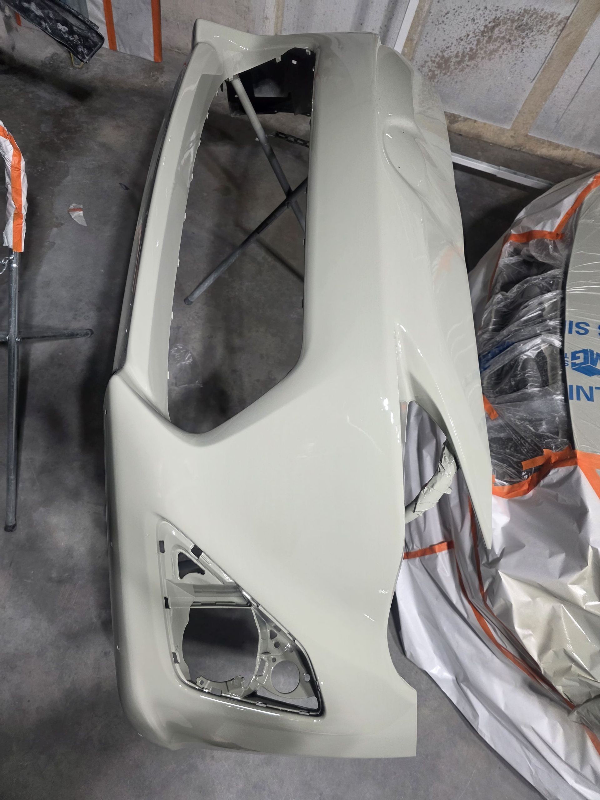 White car bumper resting on the floor in a workshop, with a headlight opening and masking plastic nearby