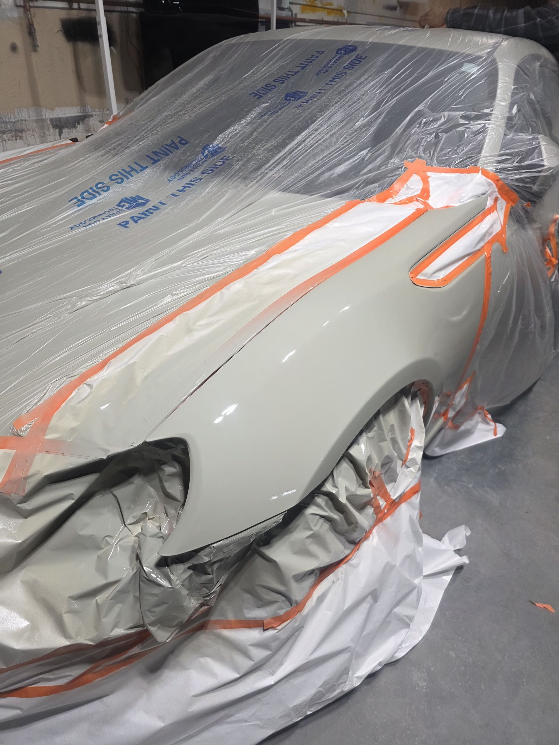 Car under repair in a body shop, wrapped in plastic and masking tape, with primer-gray paint visible