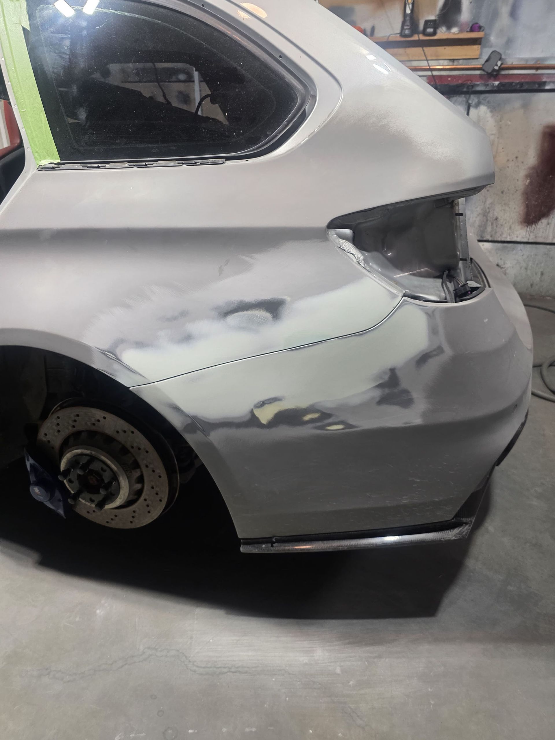 Rear side of a silver car in a garage, with damaged body panels and a removed rear wheel.