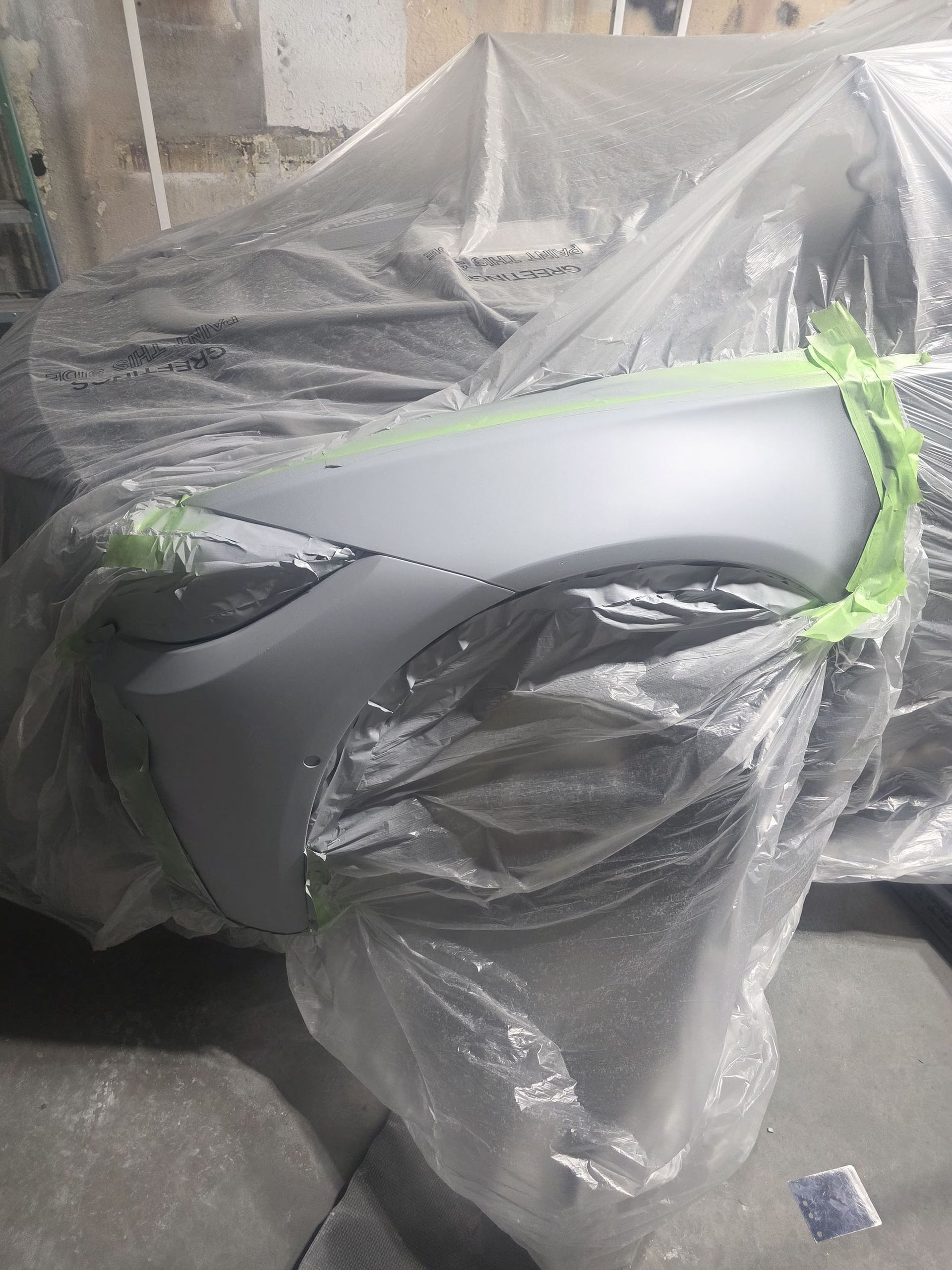 Covered gray car part wrapped in plastic and green tape in a workshop or garage