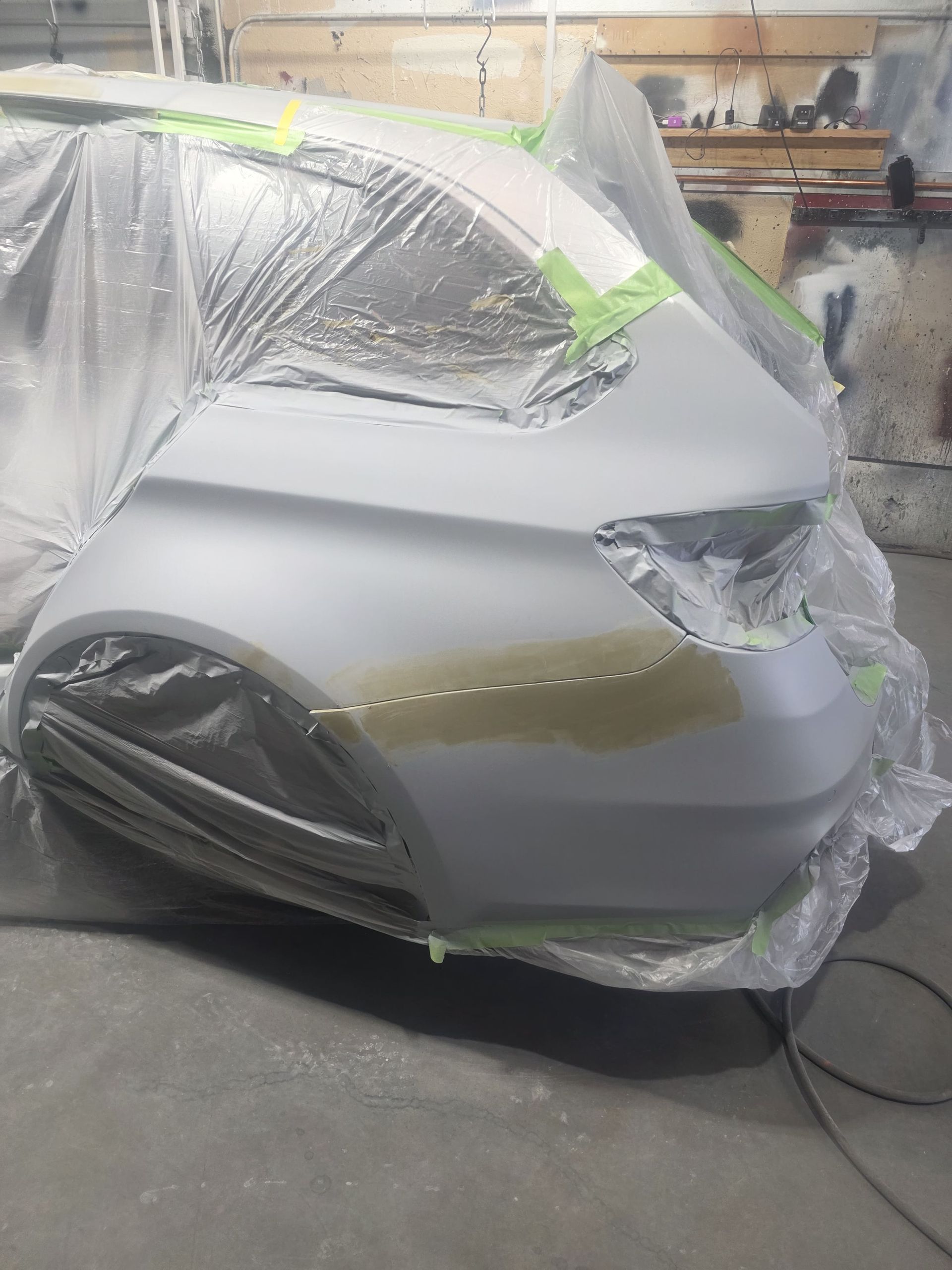 Car wrapped in masking plastic and tape in a paint shop, primed gray with exposed wheels.