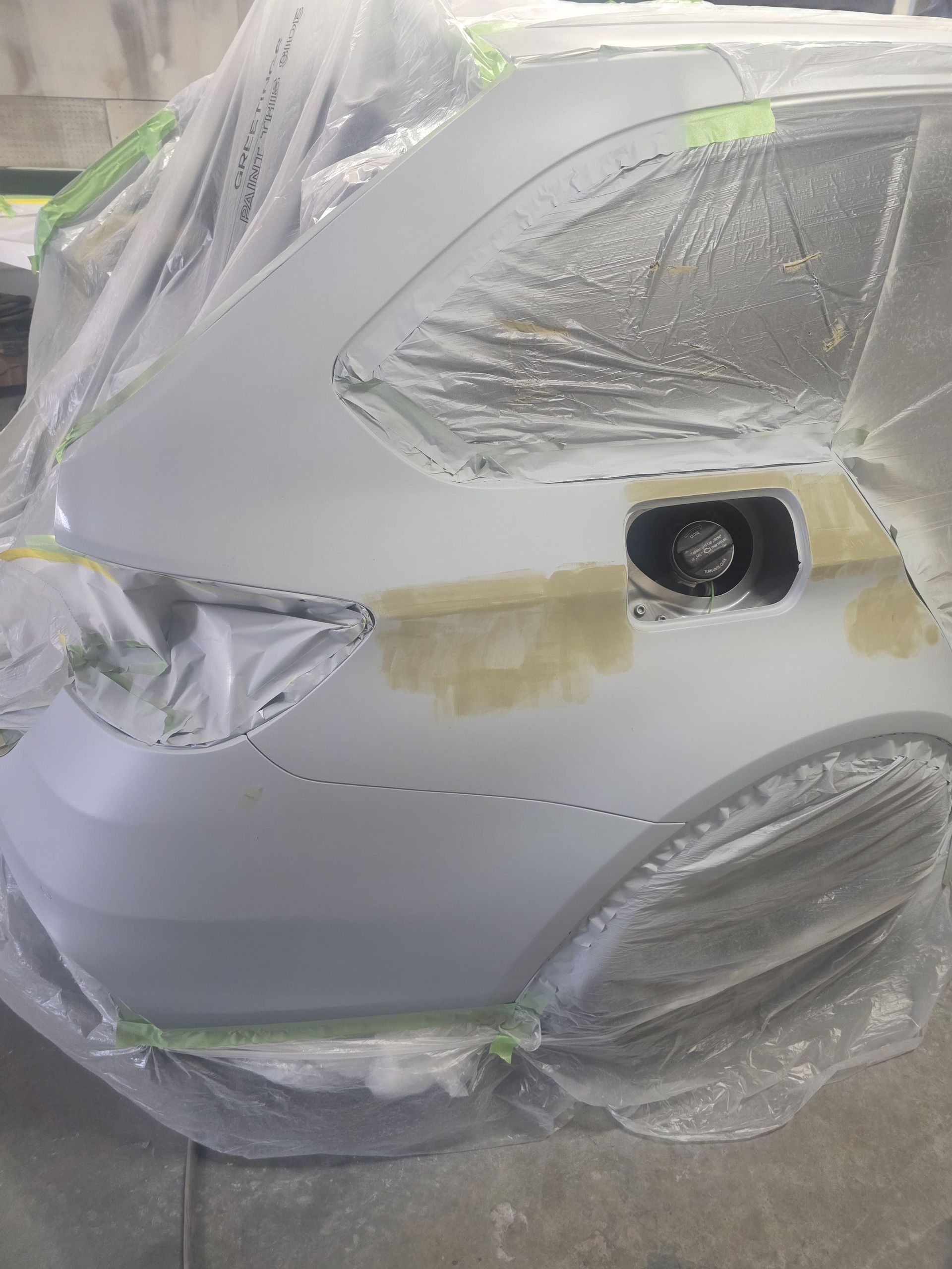 Car front masked with plastic and tape, showing taped-off headlight and body panels during paint prep