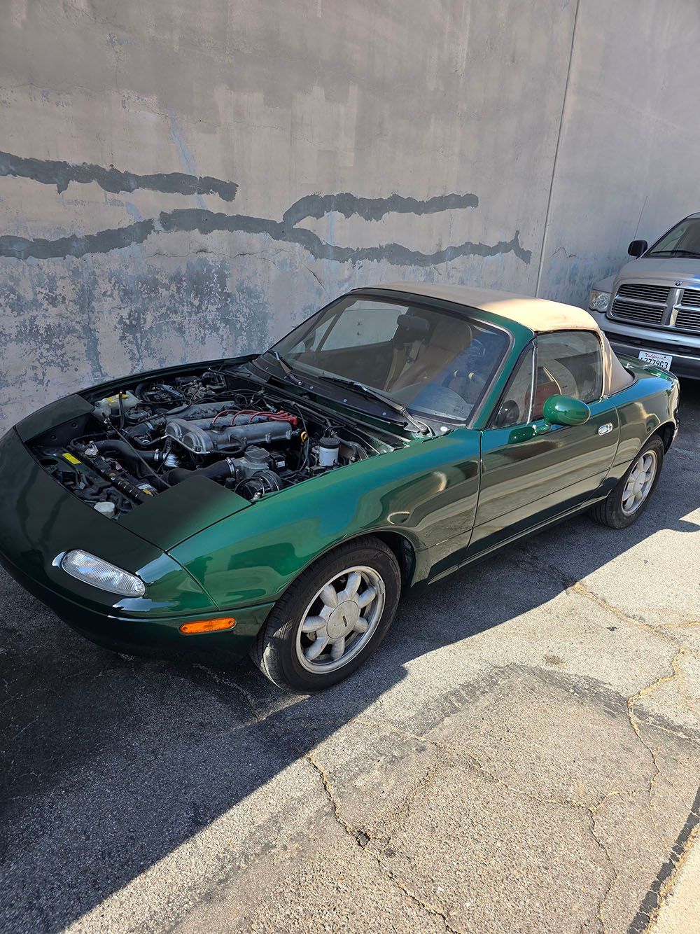 Green convertible sports car with hood open, parked beside a concrete wall in sunlight.