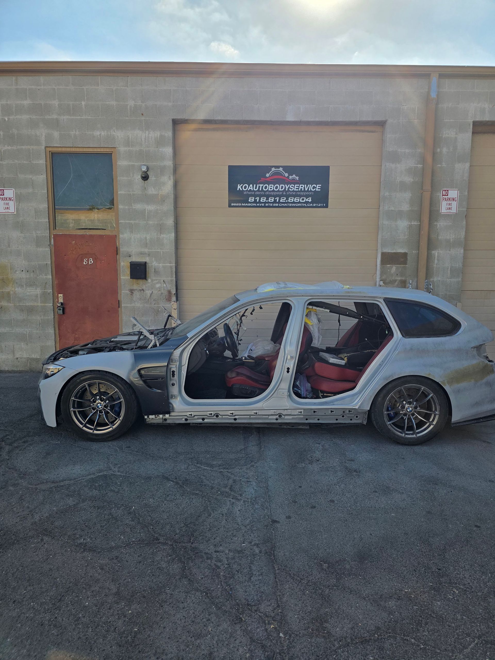 Silver hatchback with missing body panels parked in front of a garage door and red entrance door