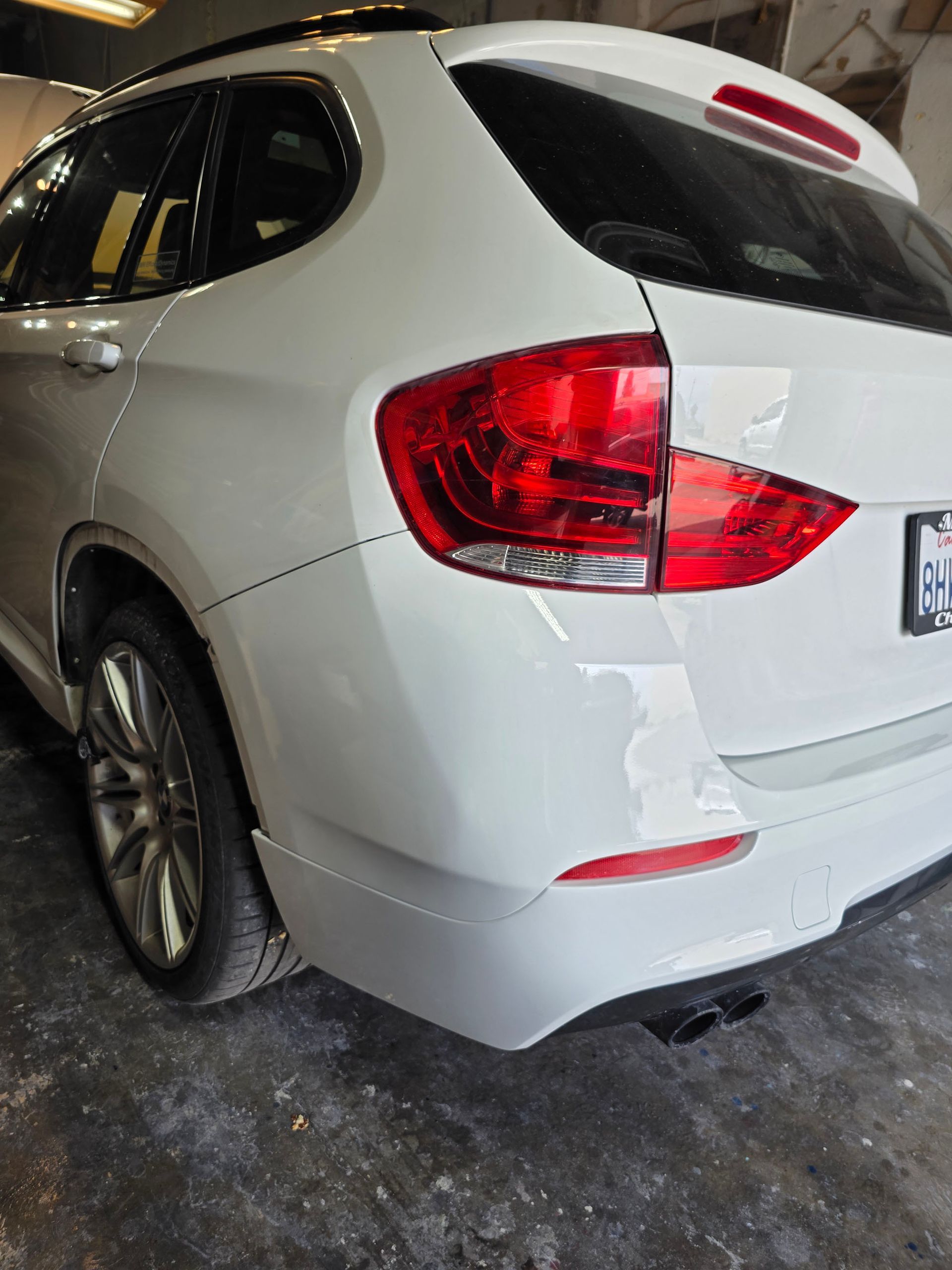 White SUV parked in a garage, rear-left view, with red taillights and a visible rear bumper scrape