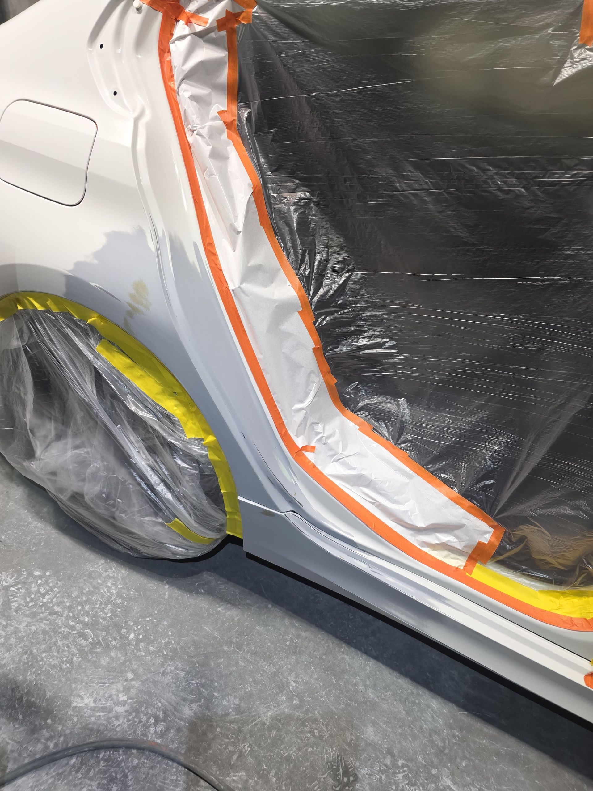 Car with black plastic wrap and orange tape over body panels, silver wheel visible in a garage-like setting
