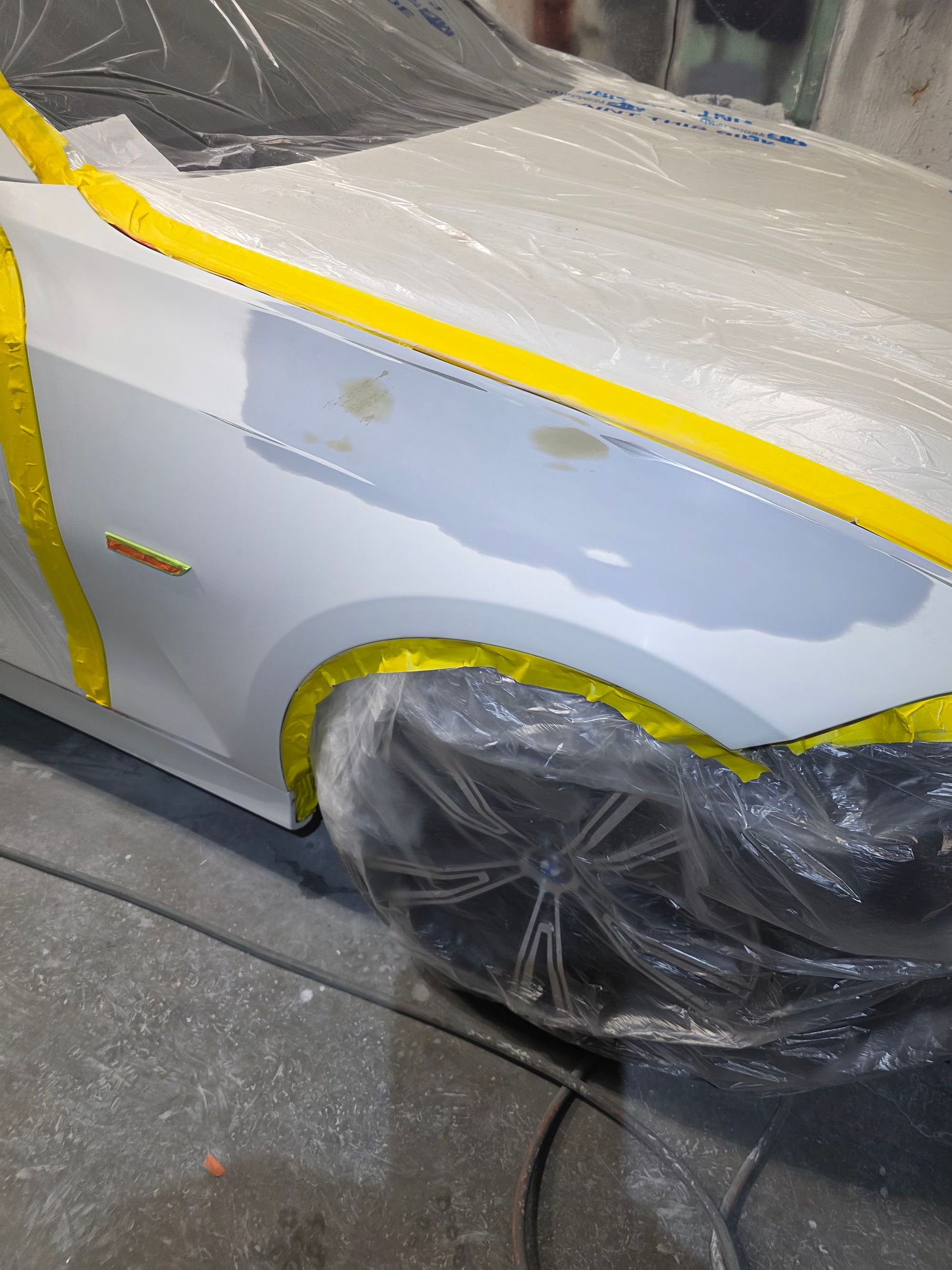 White car body being repainted with yellow masking tape and plastic covering the wheels
