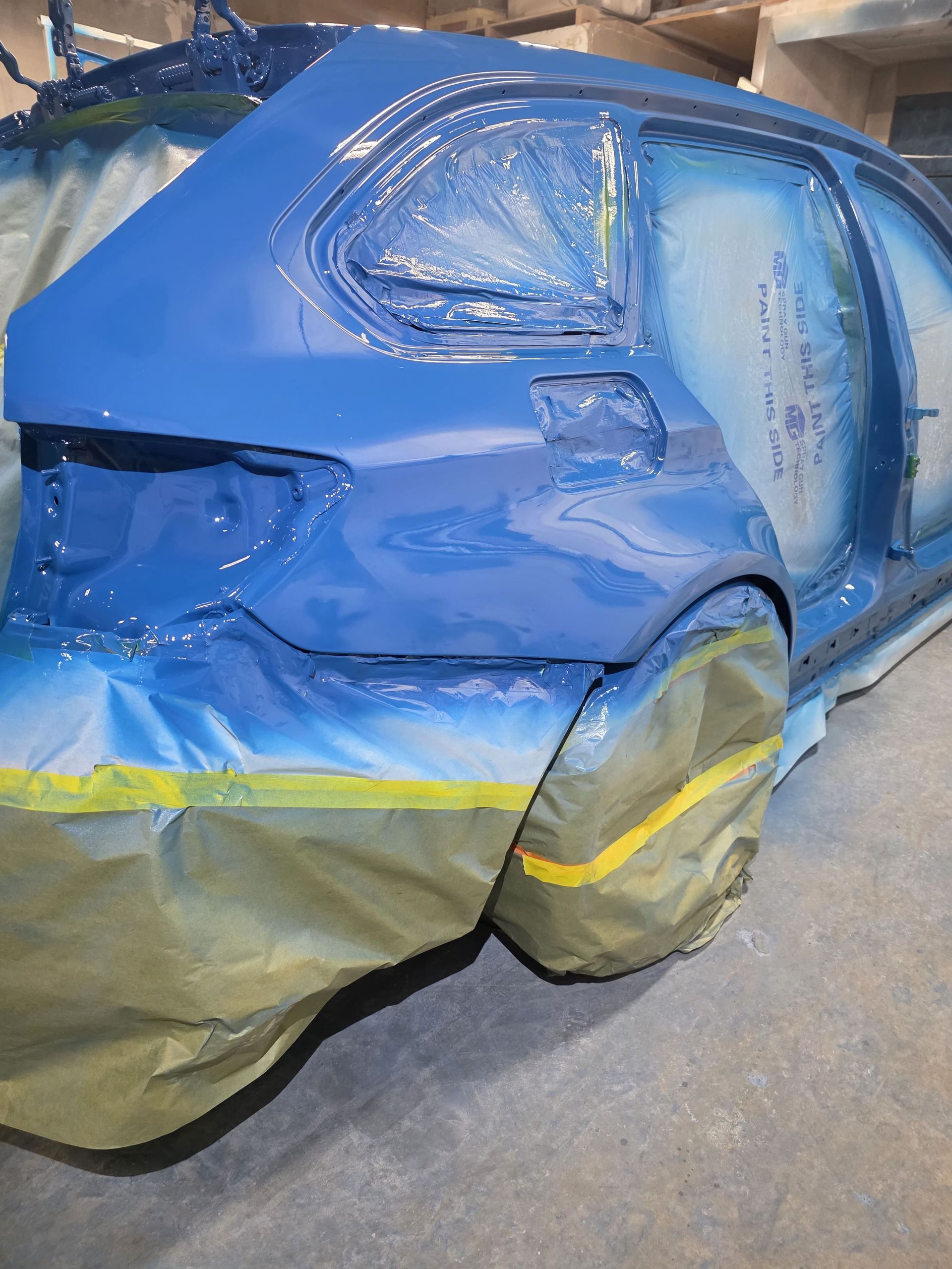 Blue car body in a paint booth, partially masked with tape and paper during painting or repair.