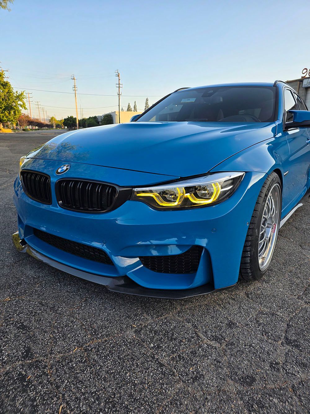 Blue BMW sports car with yellow headlights parked on gravel at sunset
