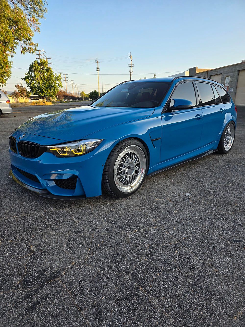Blue sports sedan parked on gravel, low angle view with trees and industrial buildings in the background