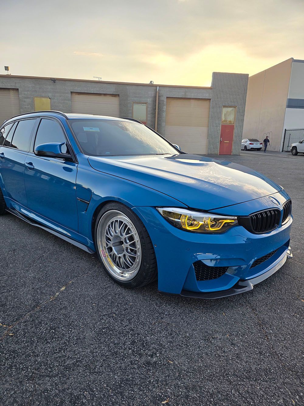 Blue sports sedan parked on gravel in front of an industrial building at sunset