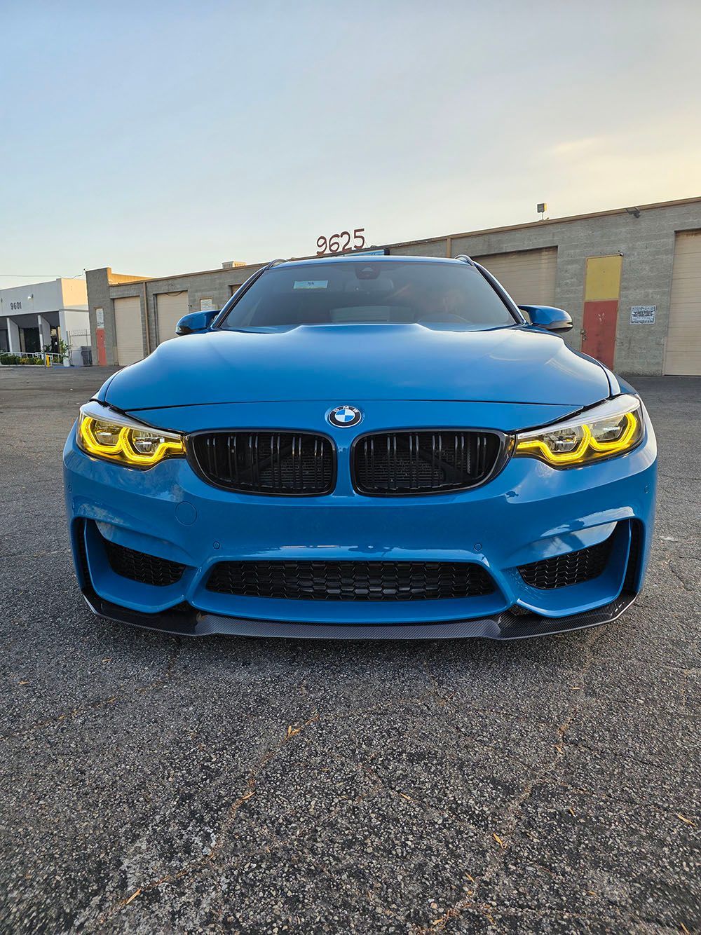 Blue BMW front view with yellow headlights parked on pavement near industrial buildings at dusk