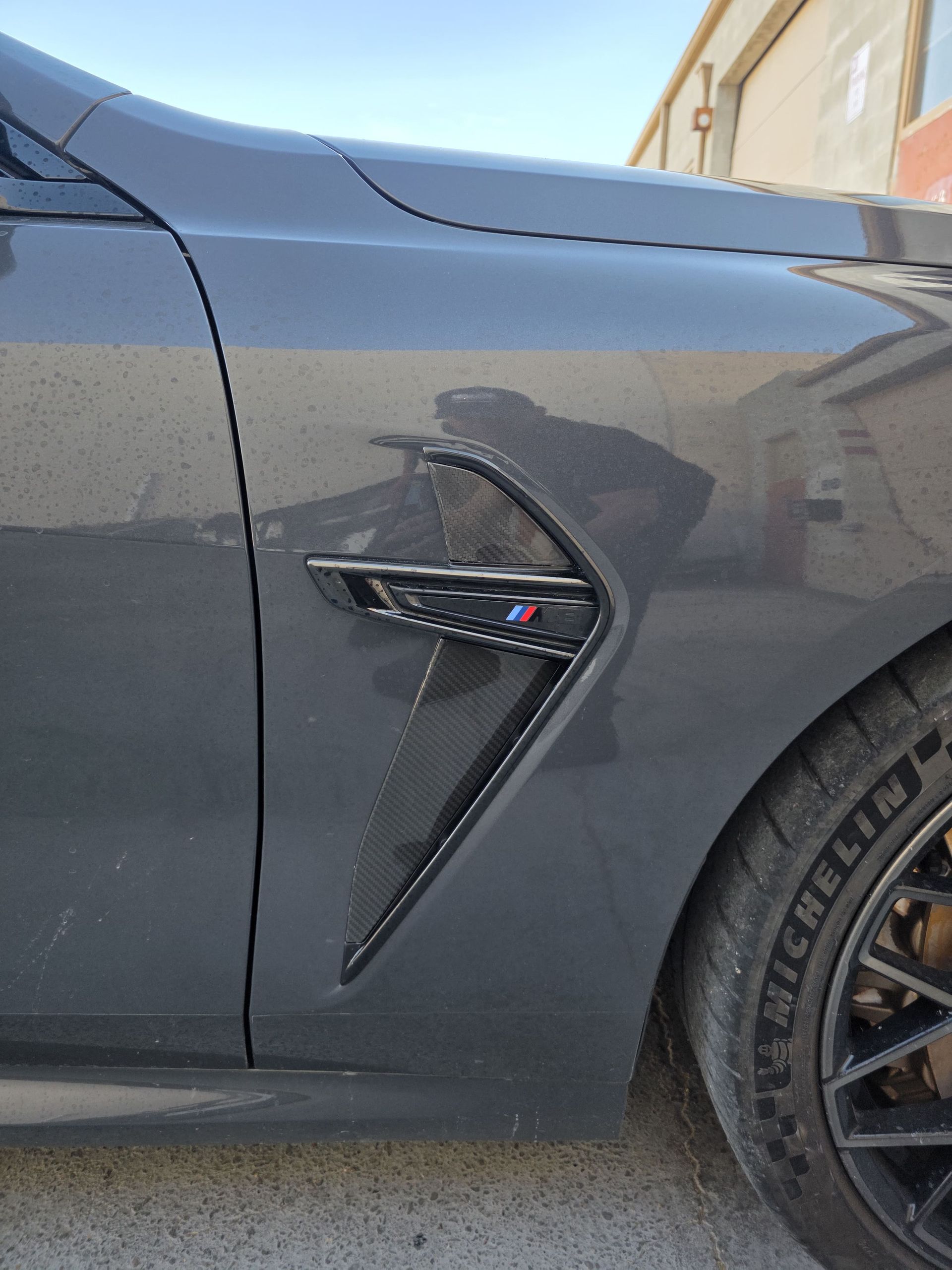 Close-up of a dark gray car’s front side panel with a vent detail and front wheel.