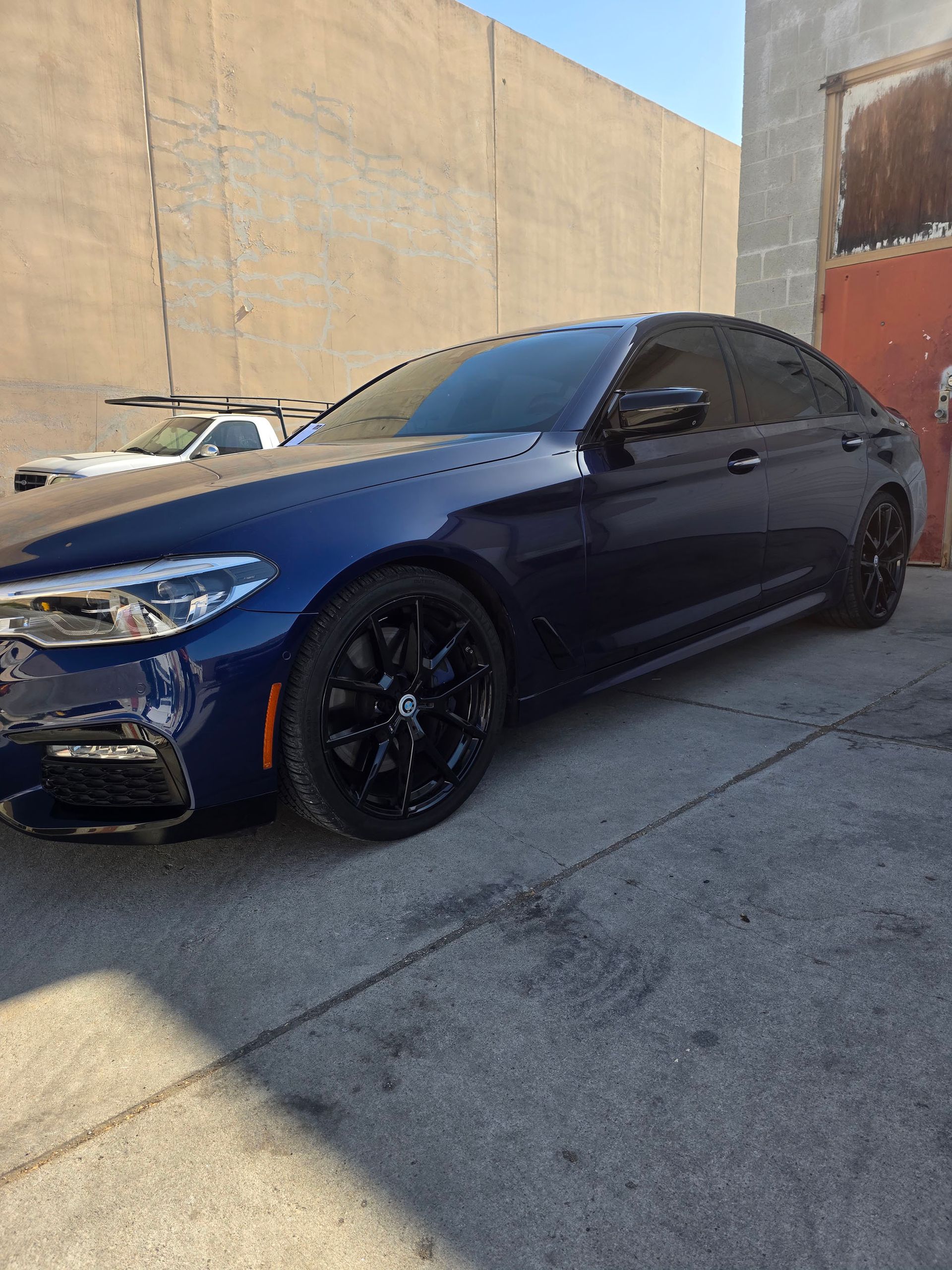 Dark blue sedan with black rims parked in an alley beside a tan wall under bright sunlight