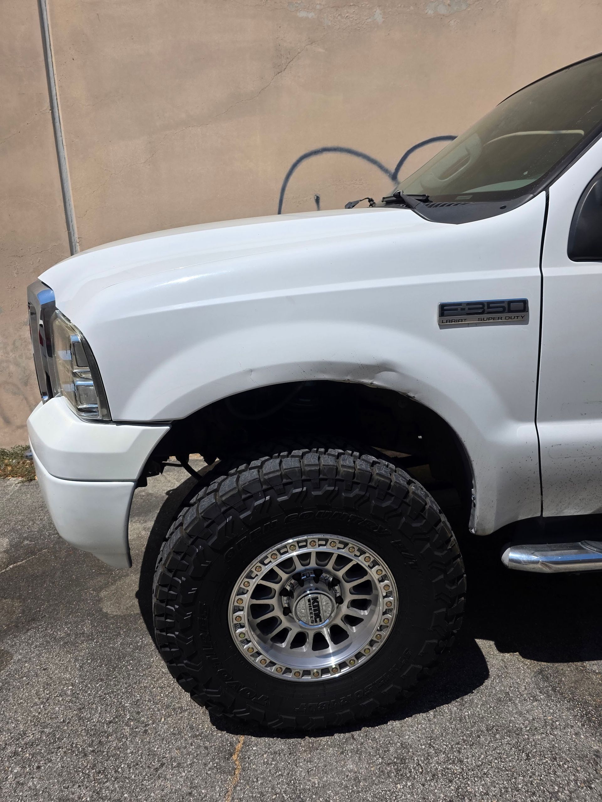 White pickup truck with a lifted front wheel and large off-road tire parked by a tan wall.