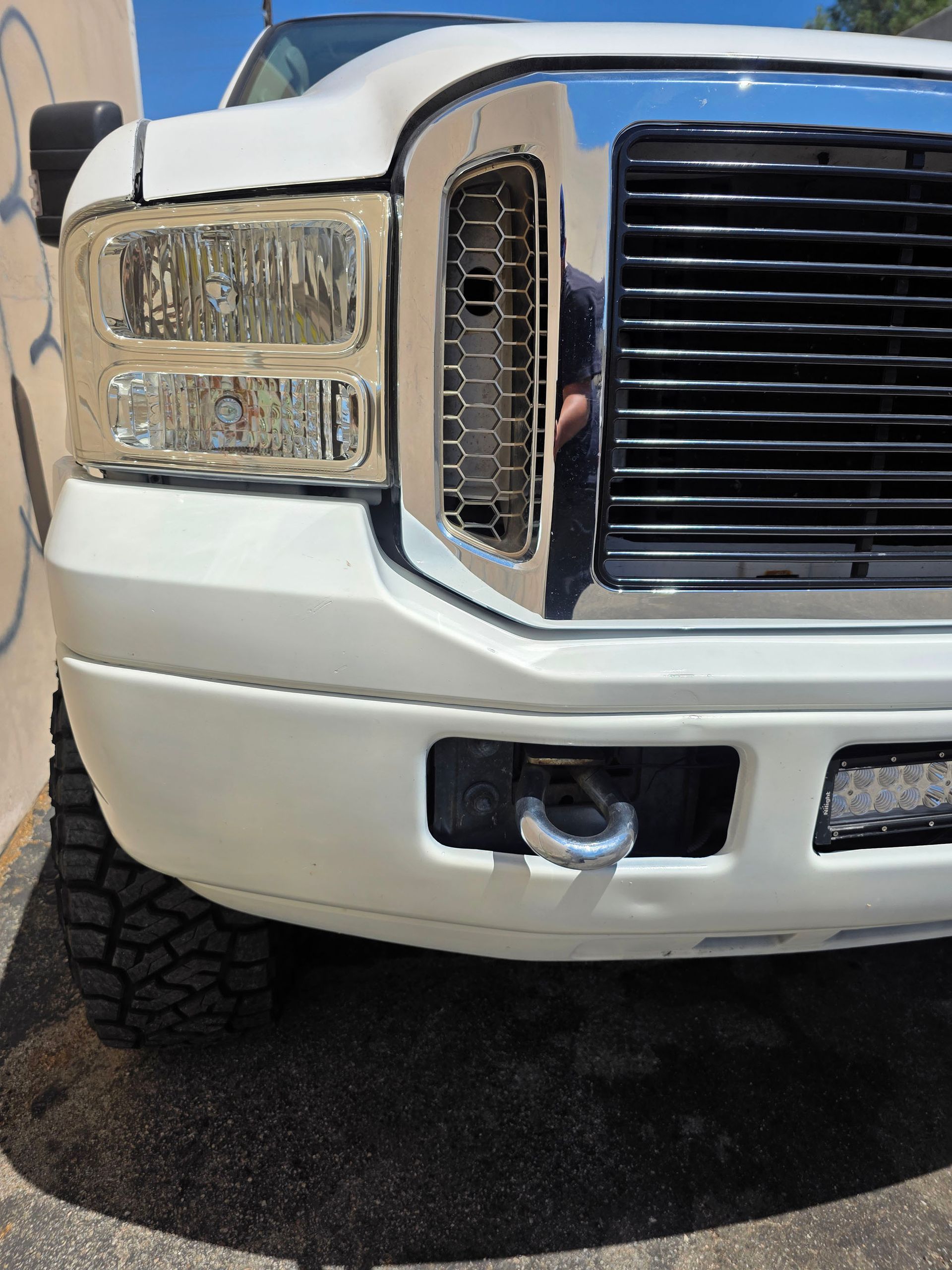Close-up of a white pickup truck’s front grille and bumper with a tow hook and off-road tires