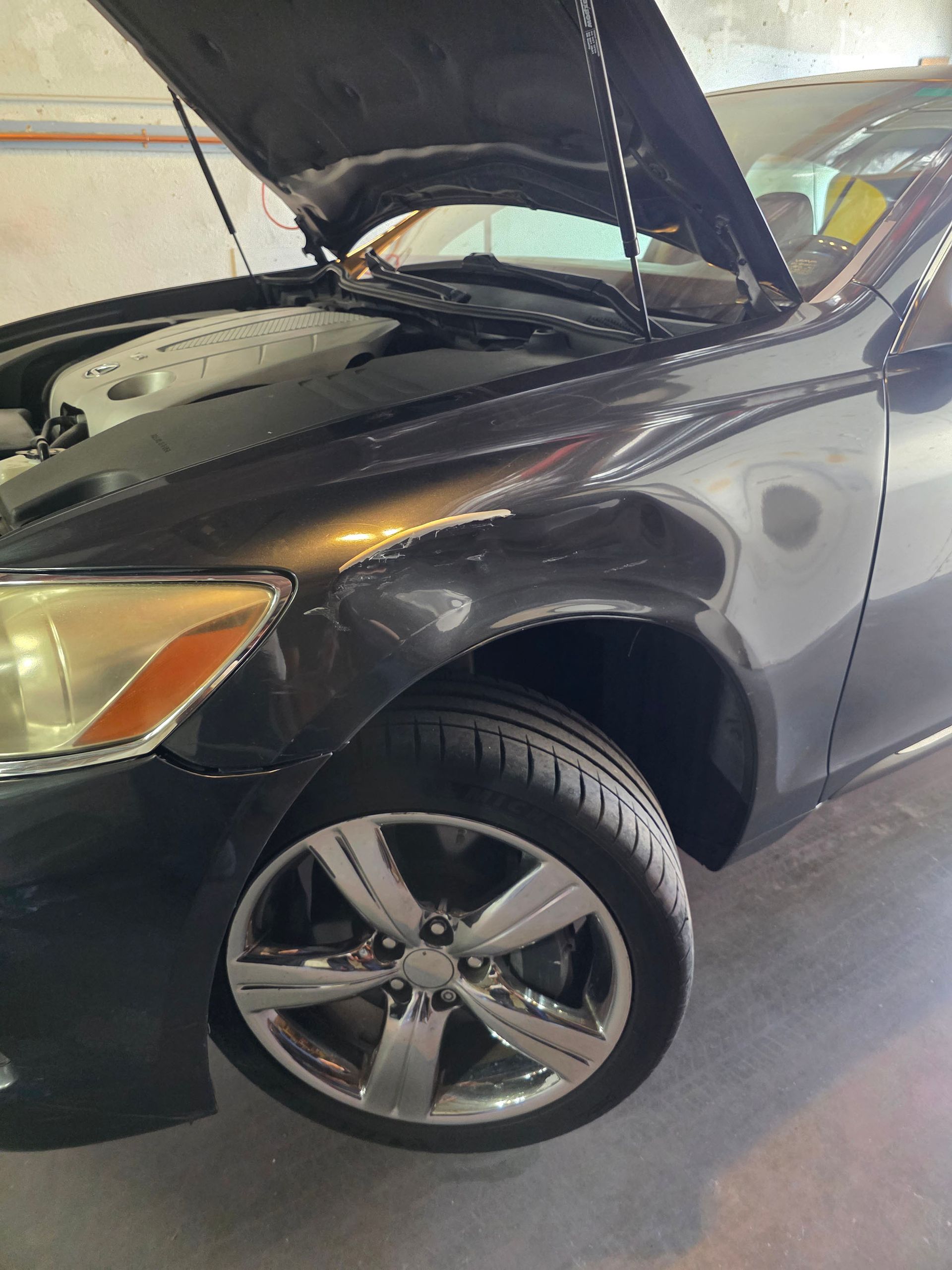 Black car in a garage with hood open, front left wheel turned outward.