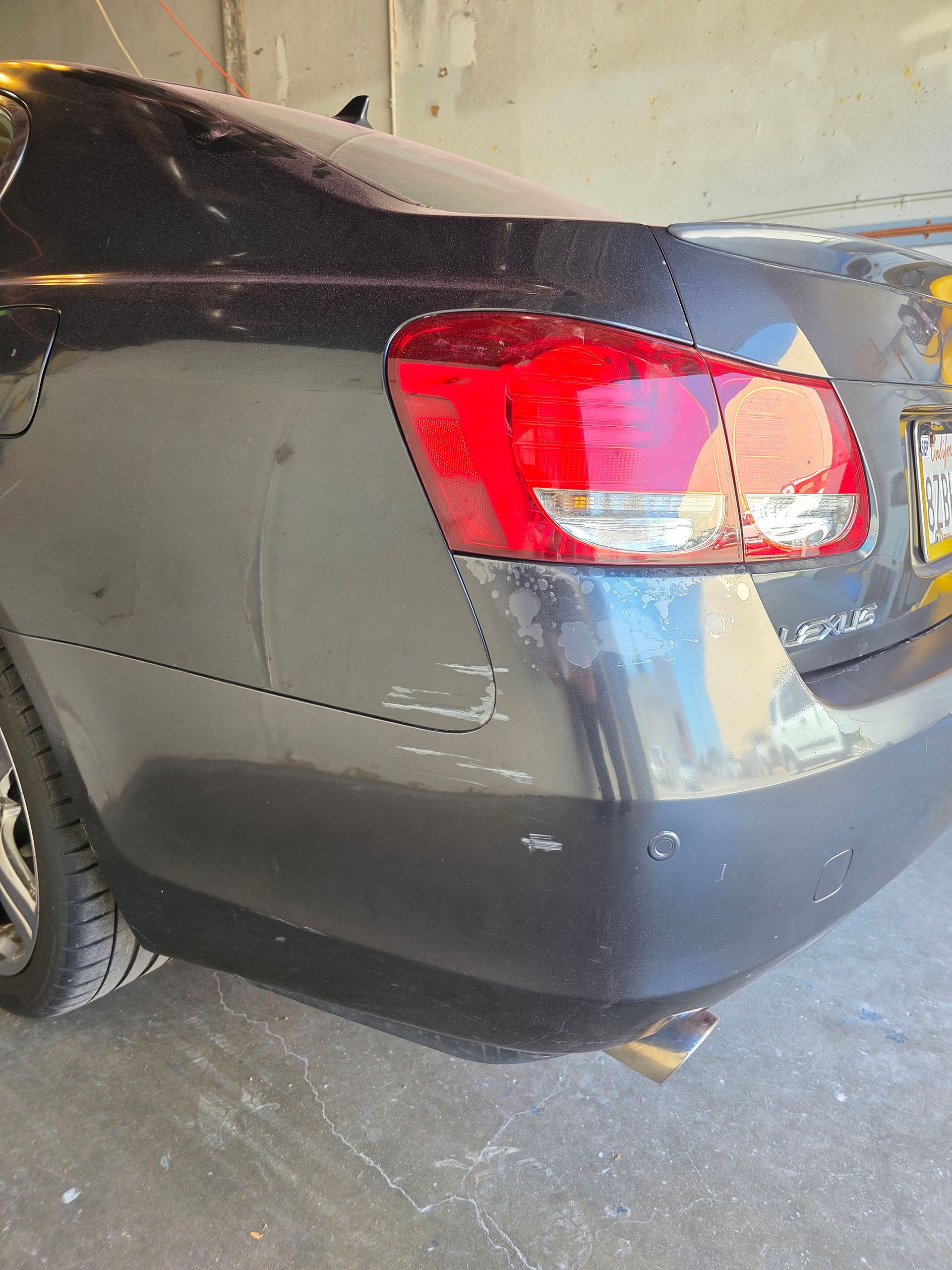 Rear corner of a dark sedan with a red taillight and visible scuff and scratch damage on the bumper and quarter panel.