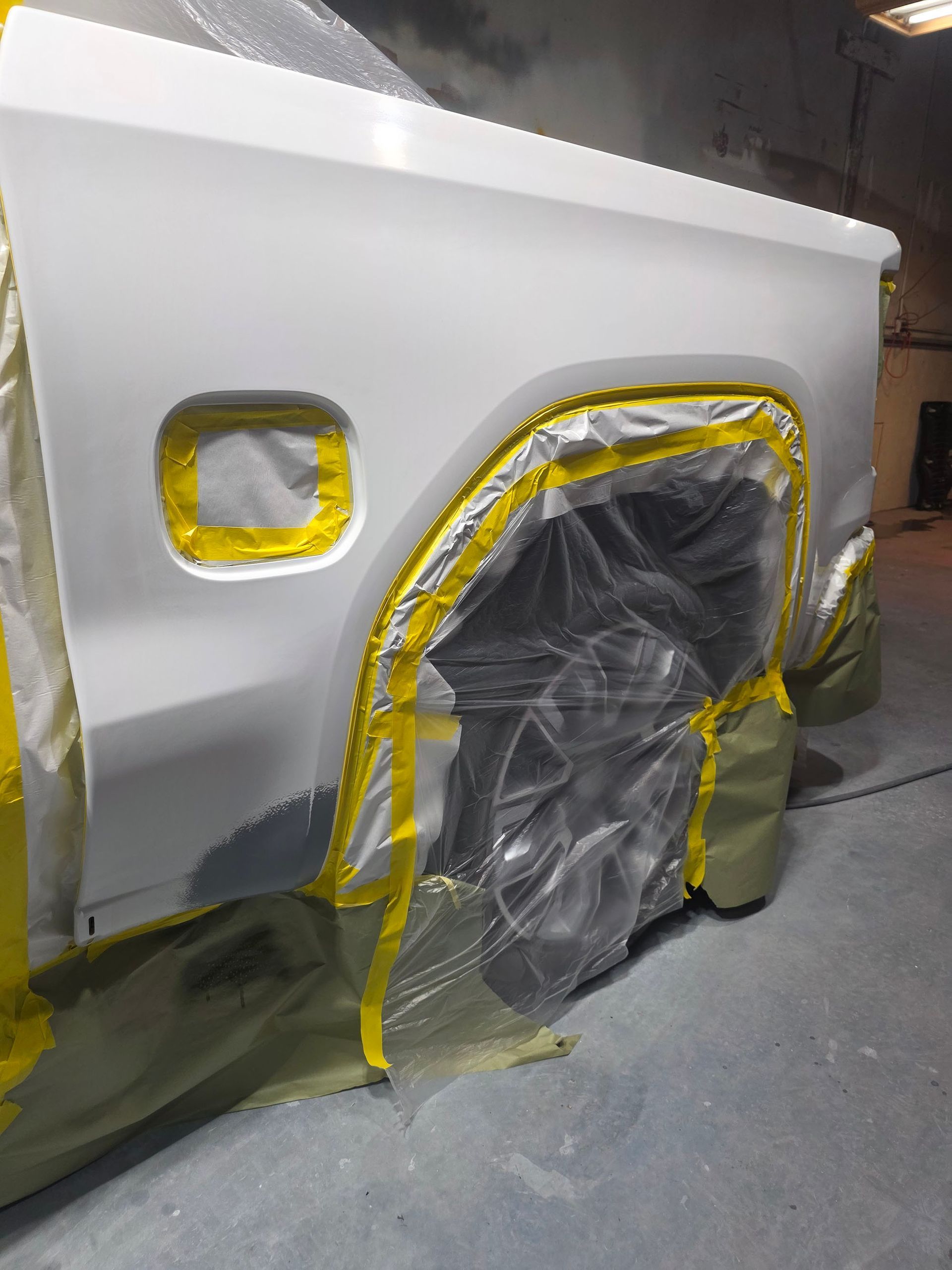 White vehicle body panel in a workshop, masked with yellow tape and plastic over the door opening