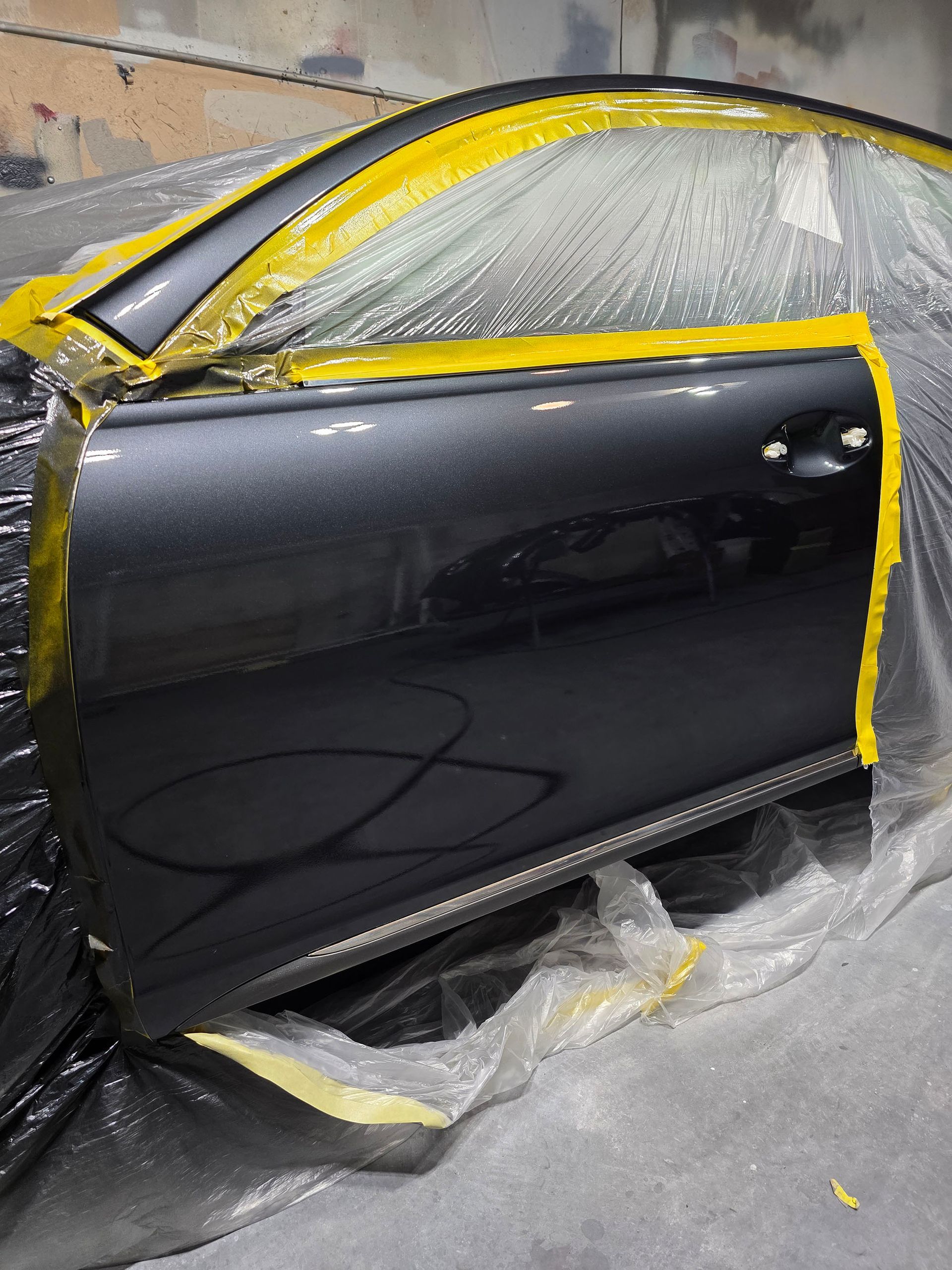 Black car door taped off and partly painted in a repair shop, covered with plastic masking.