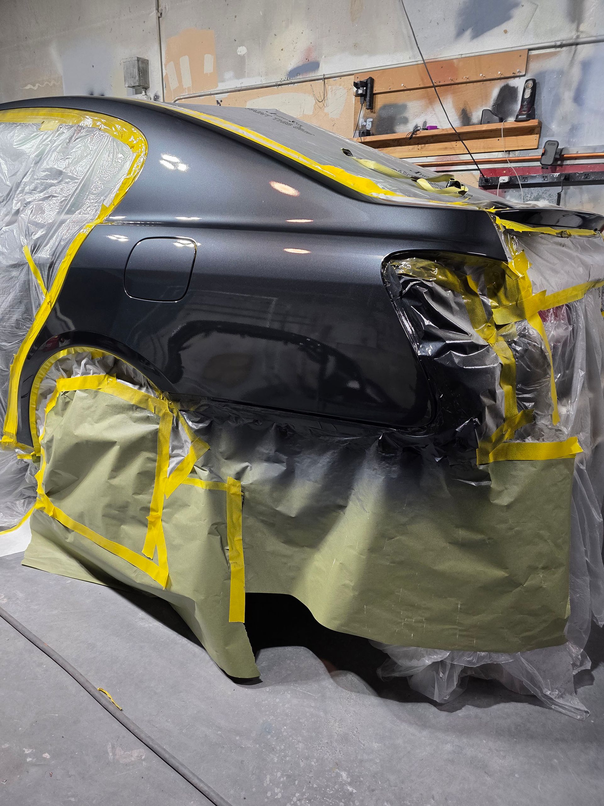 Black car body in a paint booth, masked with plastic and yellow tape during repair work