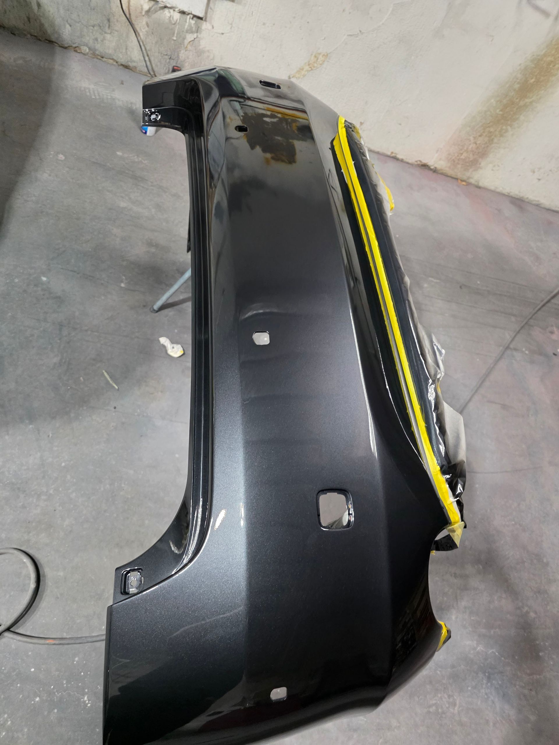 Black car bumper cover with yellow trim leaning against a wall on a concrete floor